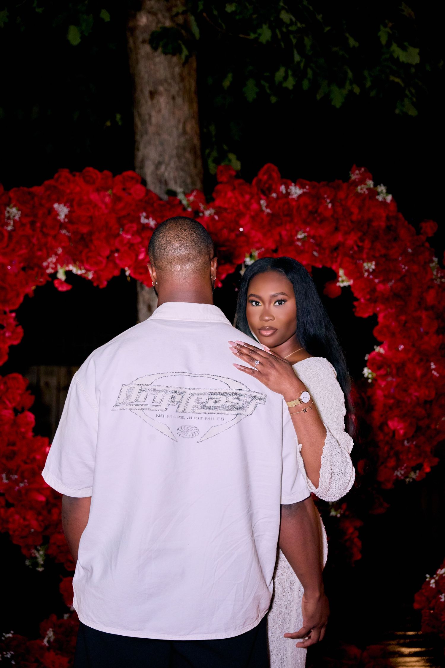 A couple poses near vibrant red flowers at night, with one person wearing a white shirt with a logo on the back.