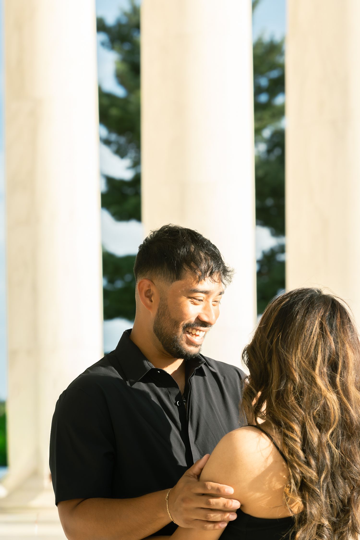 A couple shares a tender moment together while standing between white pillars on a sunny day.