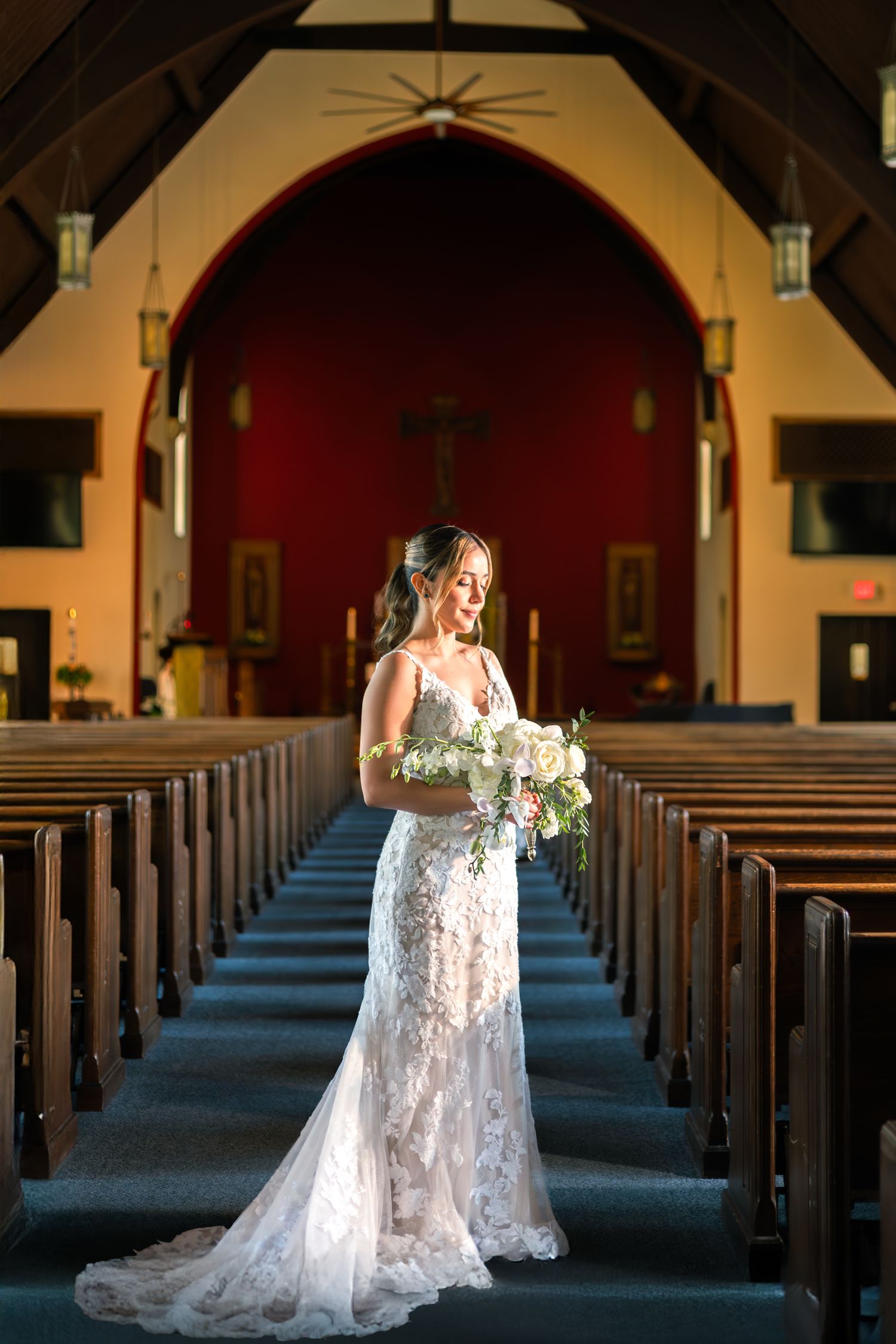 A bride in a white lace wedding dress stands in the aisle of a church with natural light streaming through the windows.