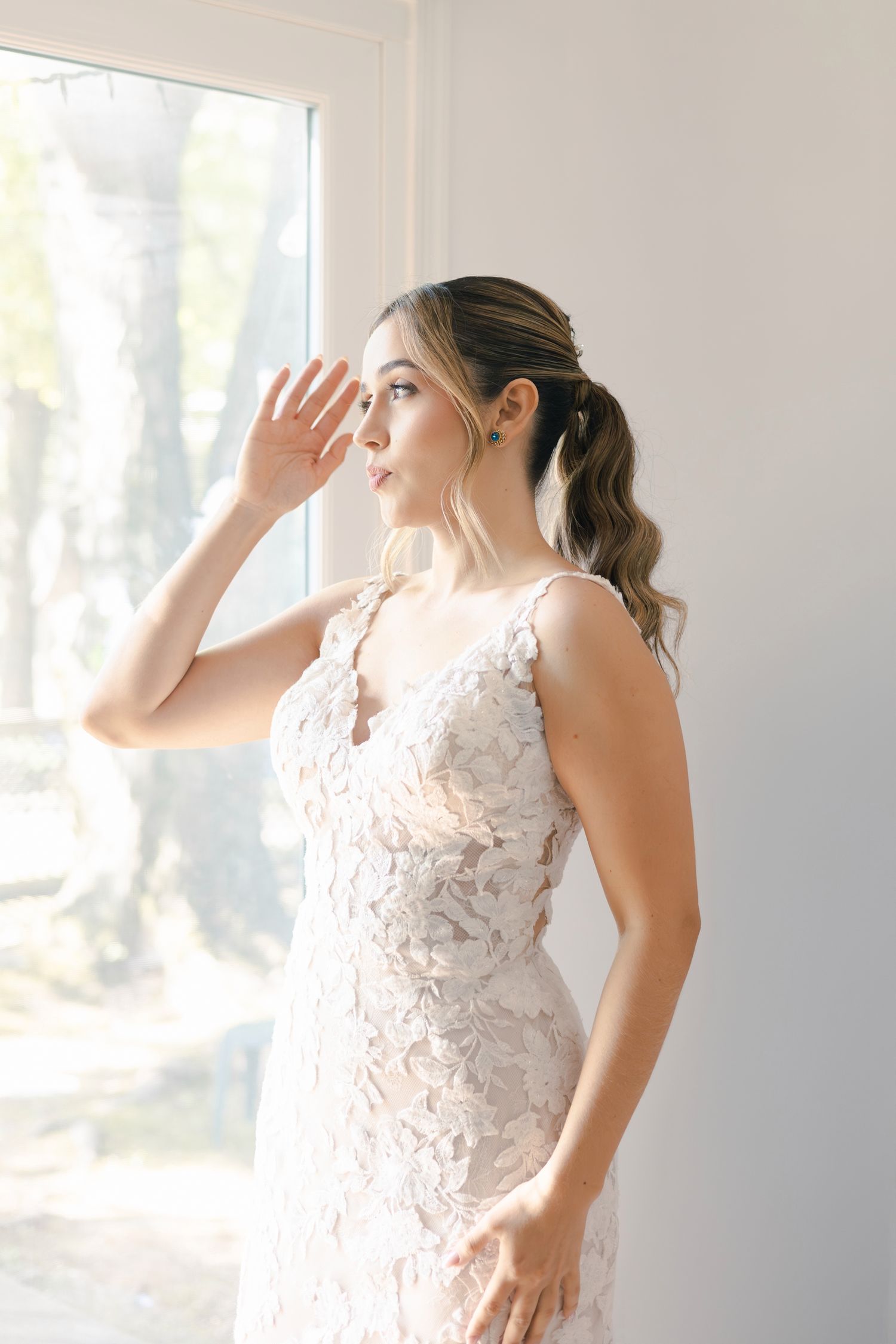 A bride in a white lace wedding dress poses gracefully in a sunlit room with a romantic ethereal atmosphere.