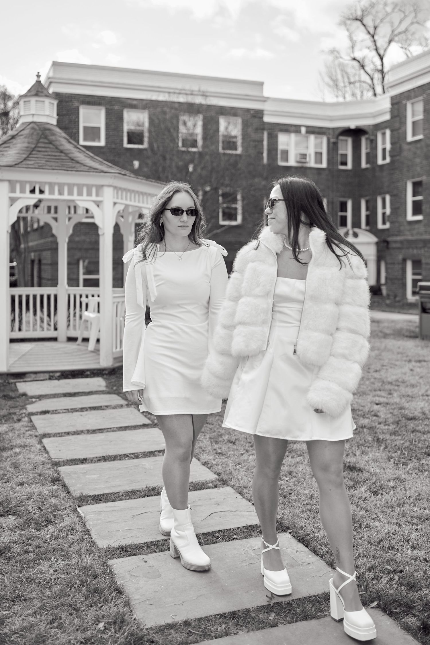 Two people in white outfits walk together on a stone path near a gazebo and brick building.