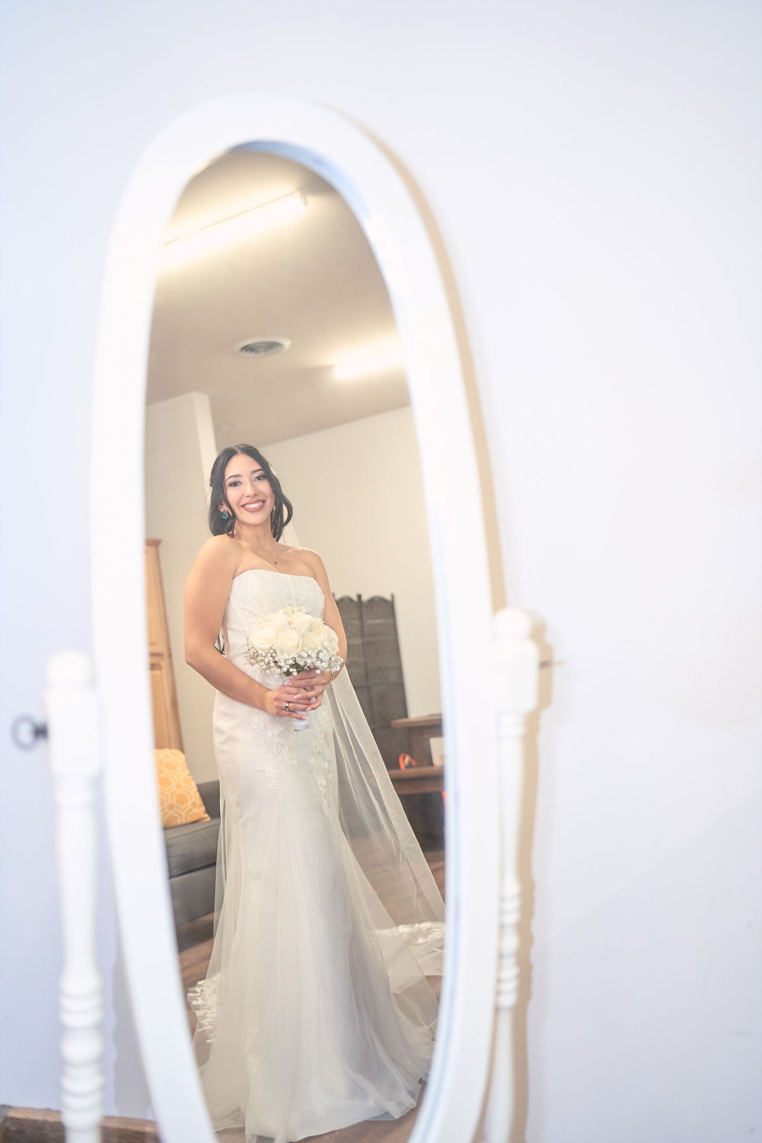 A bride in a white wedding dress looks at her reflection in an ornate full-length mirror.