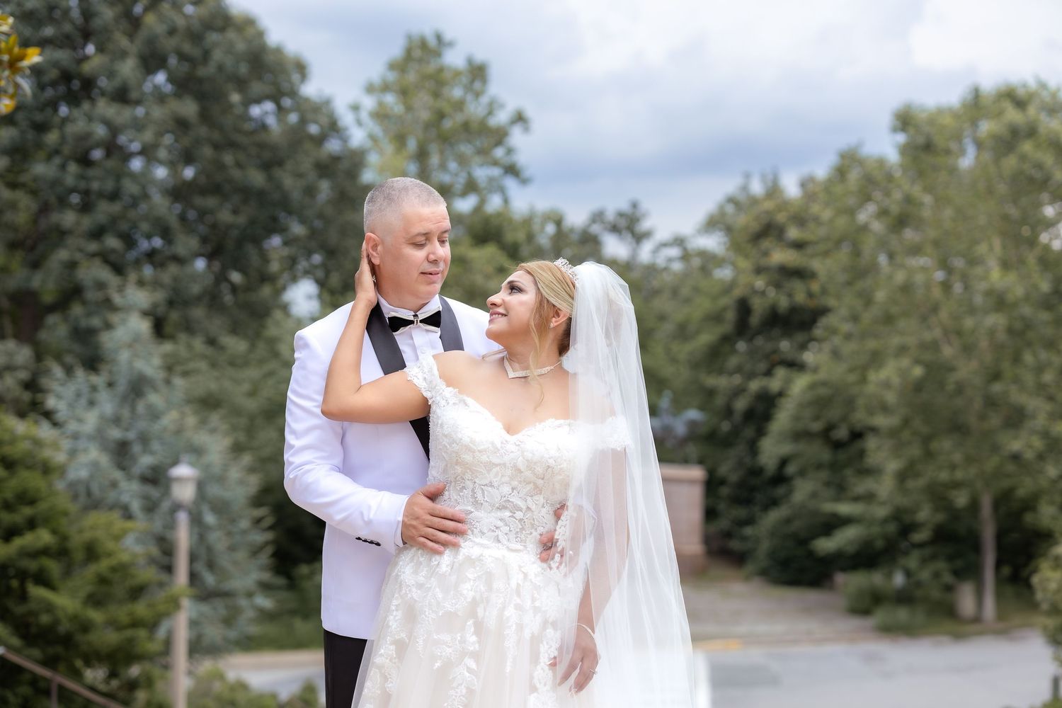 A joyful newlywed couple shares a romantic moment on a tree-lined path in their white wedding attire.