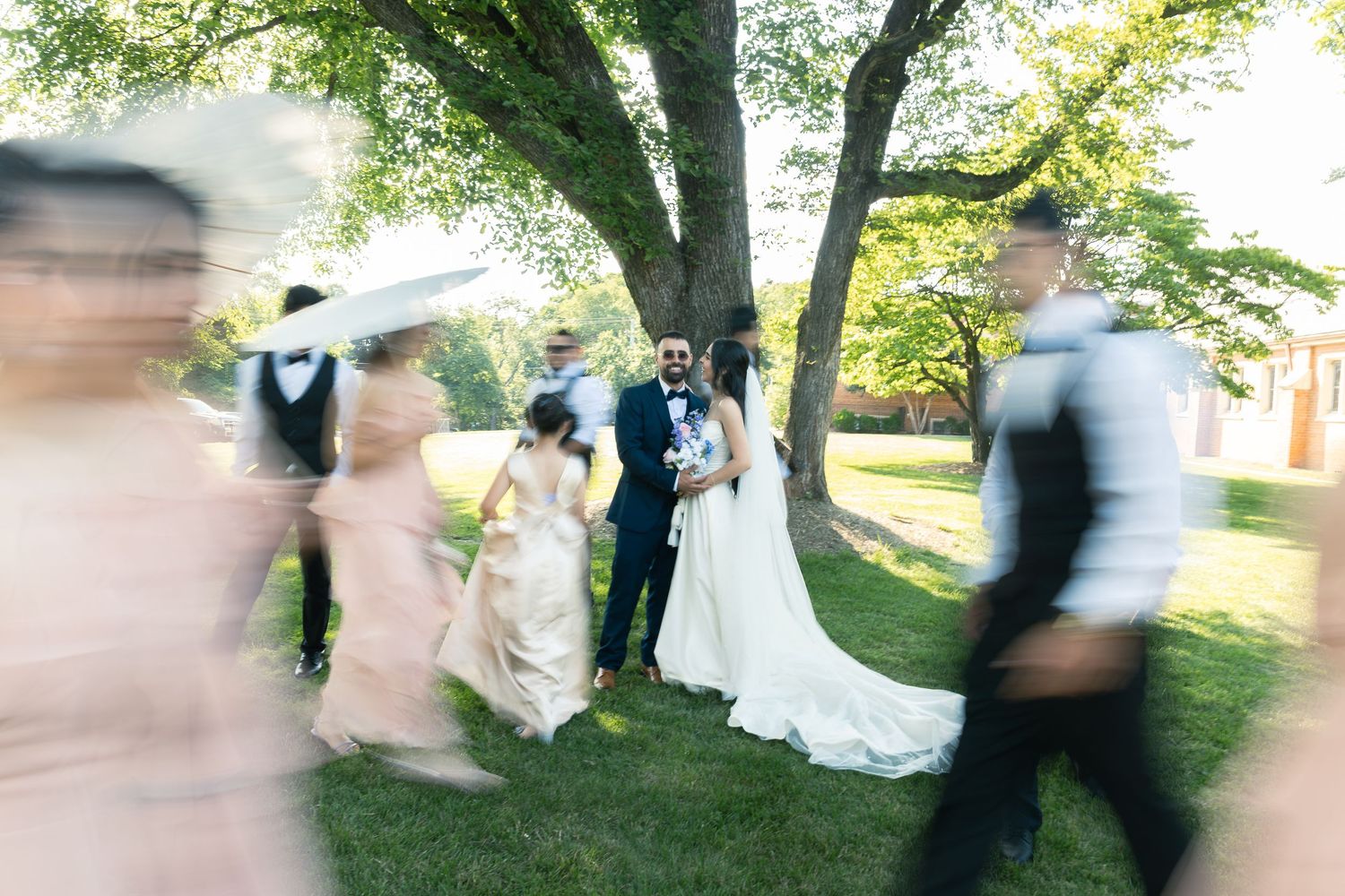 Motion blur photograph of wedding guests moving past as newlyweds stand under a tree at an outdoor ceremony.