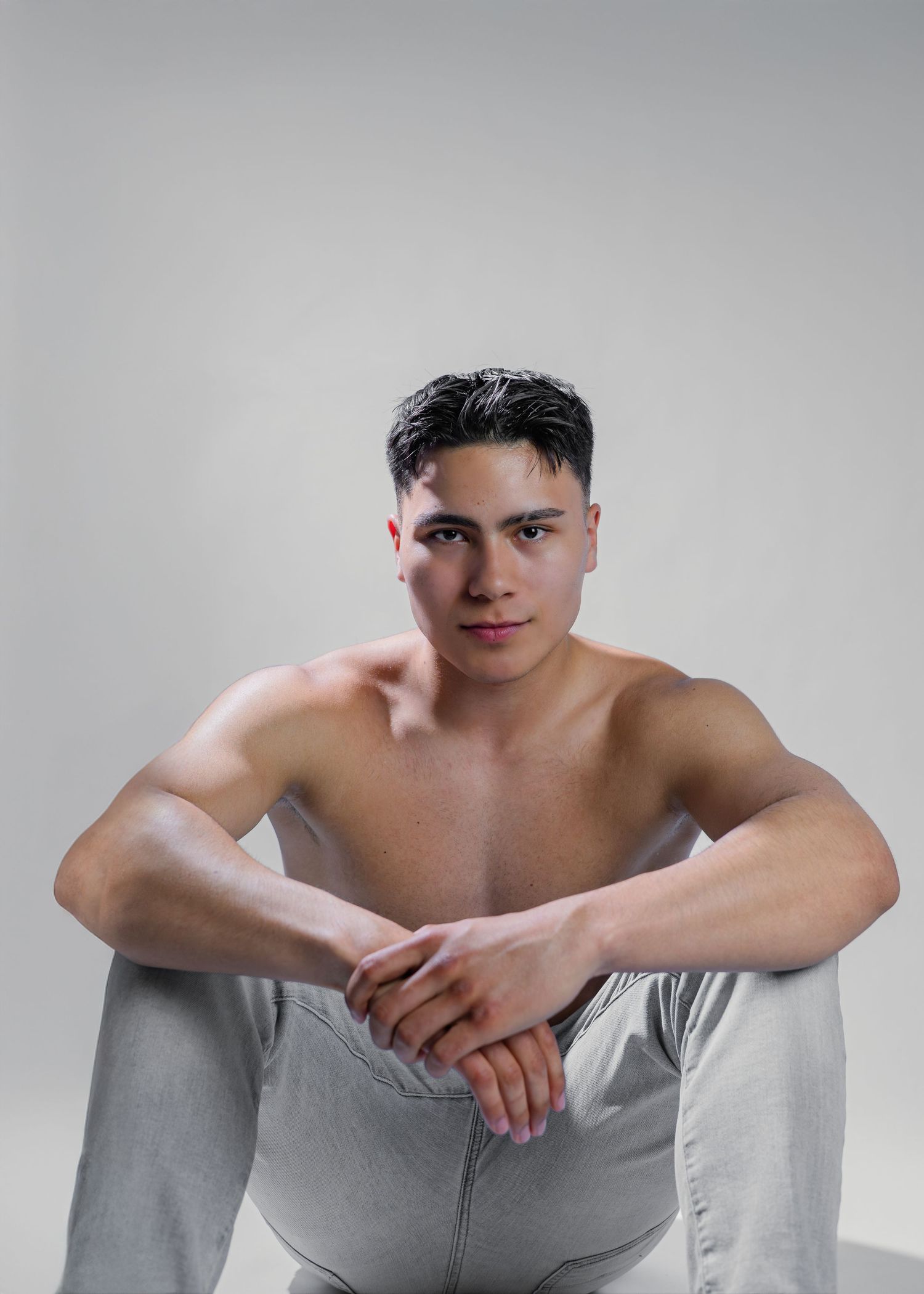 A shirtless model in light grey pants sits against a neutral grey background in a minimalist studio setting.