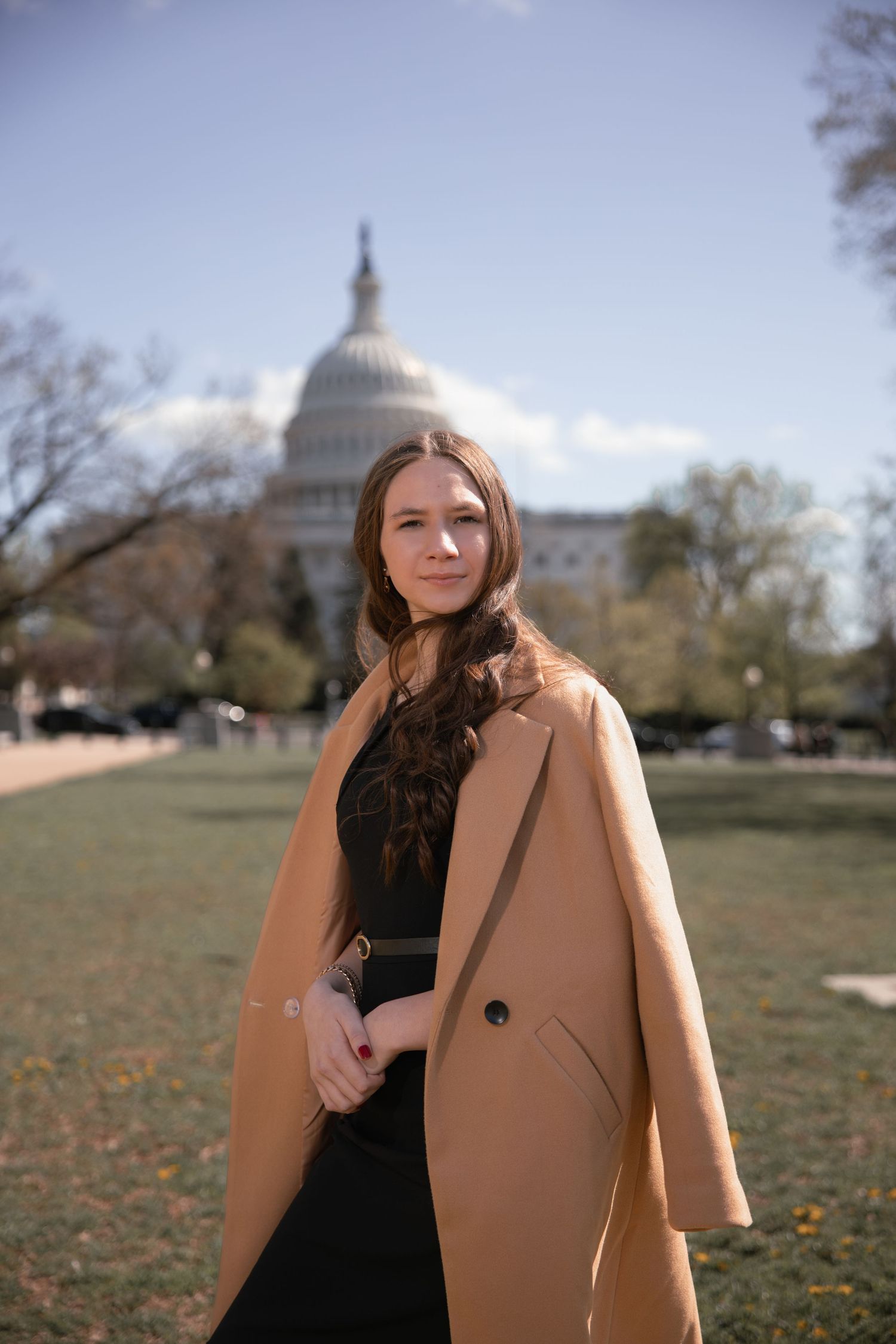 Portrait of someone in a camel coat and black outfit standing in front of the US Capitol building on a sunny day.