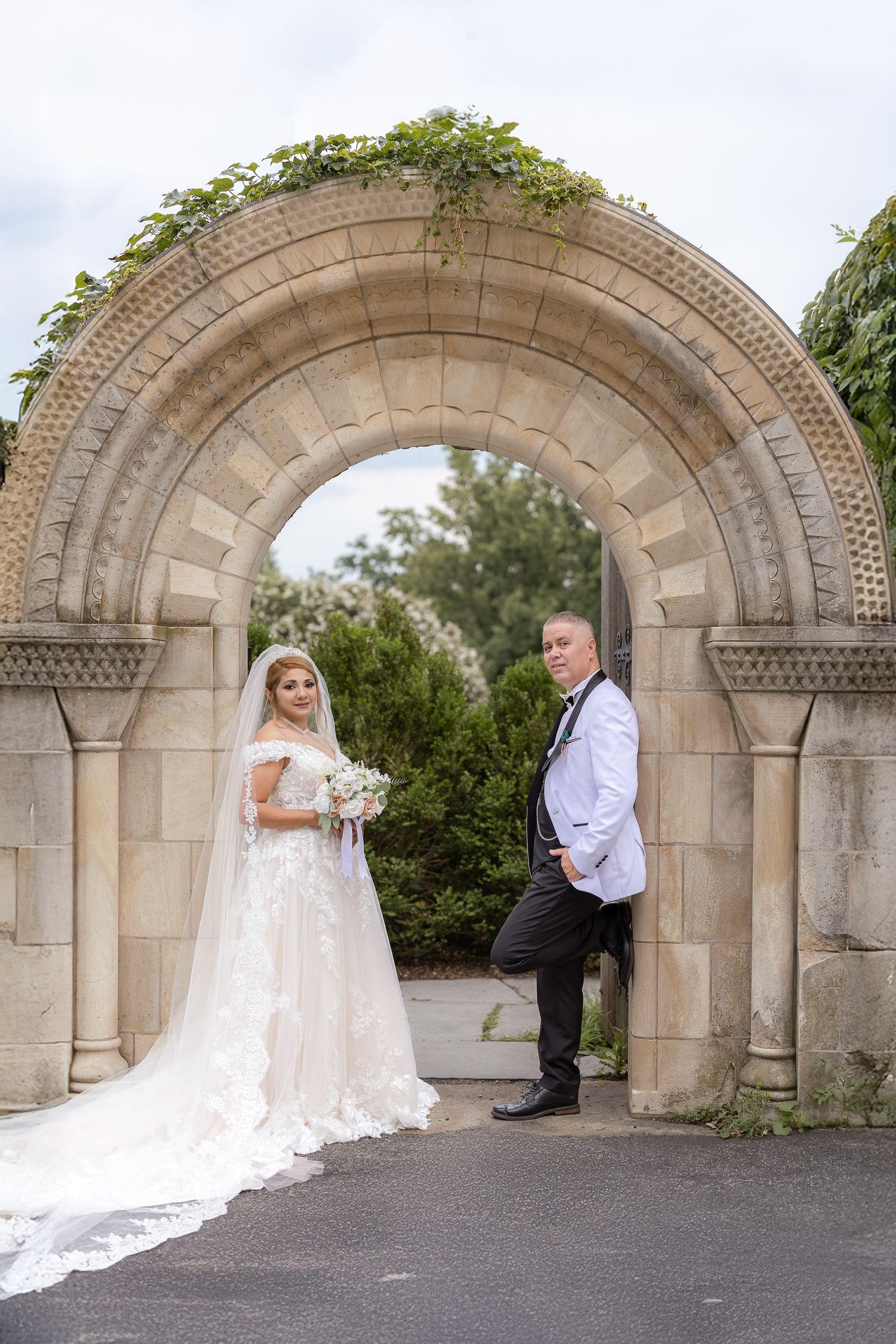 Bride and groom pose under ornate stone archway with intricate architectural details during outdoor wedding photoshoot.