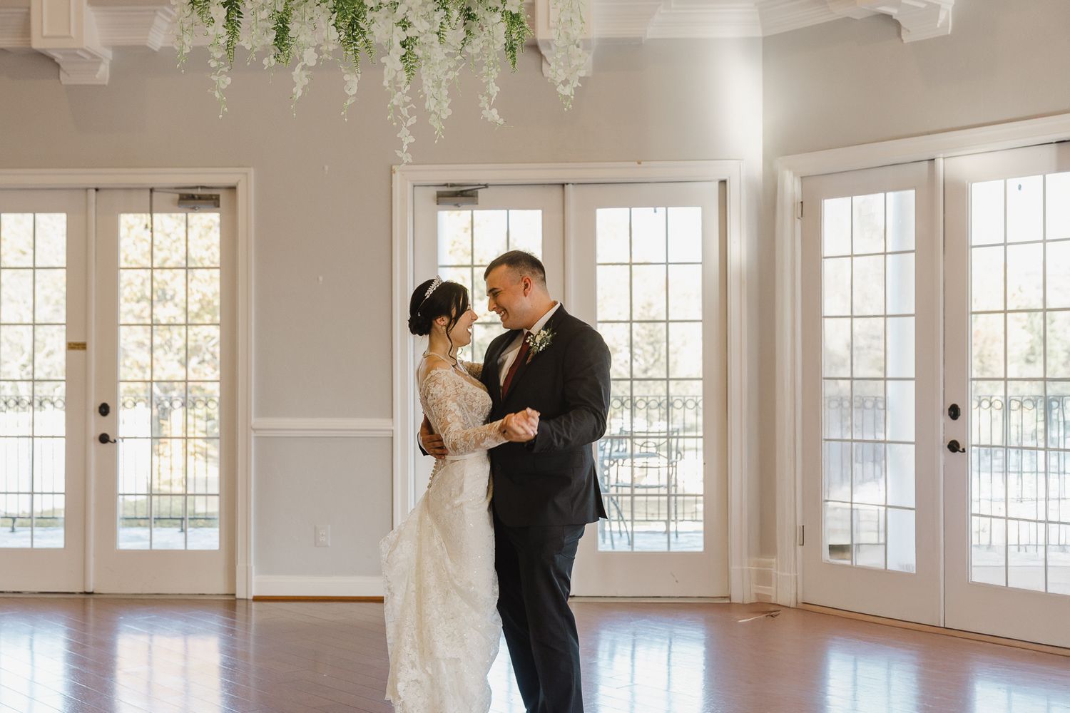 A bright white ballroom with large windows and hanging greenery showcases an elegant wedding venue setting.