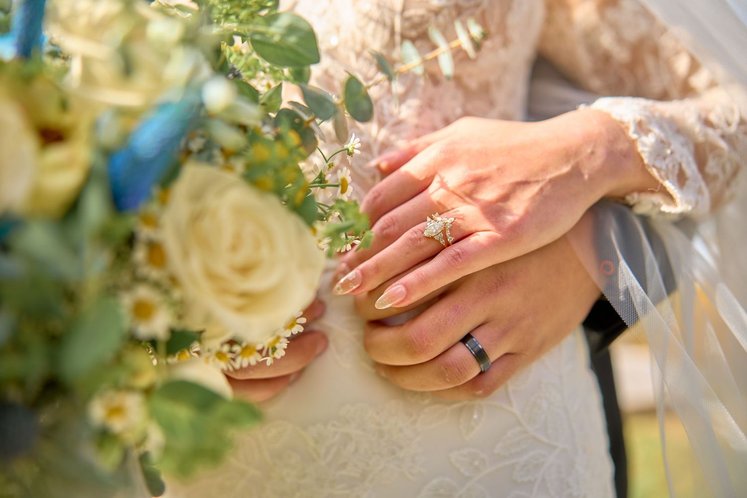 A close-up of hands wearing wedding rings next to a bouquet of cream roses and greenery.