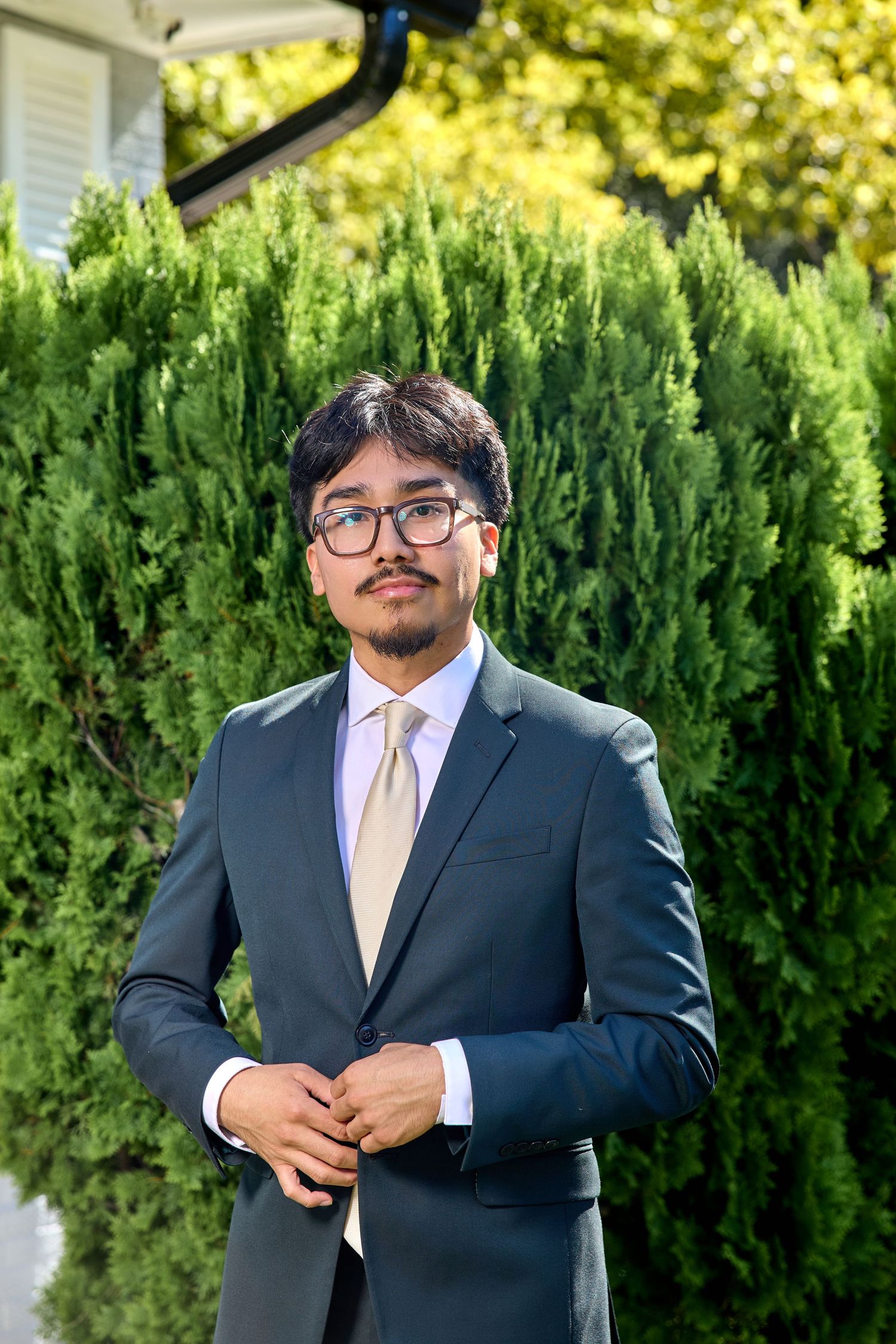 A person in a dark suit and peach tie poses outdoors against lush green shrubs in a formal setting.