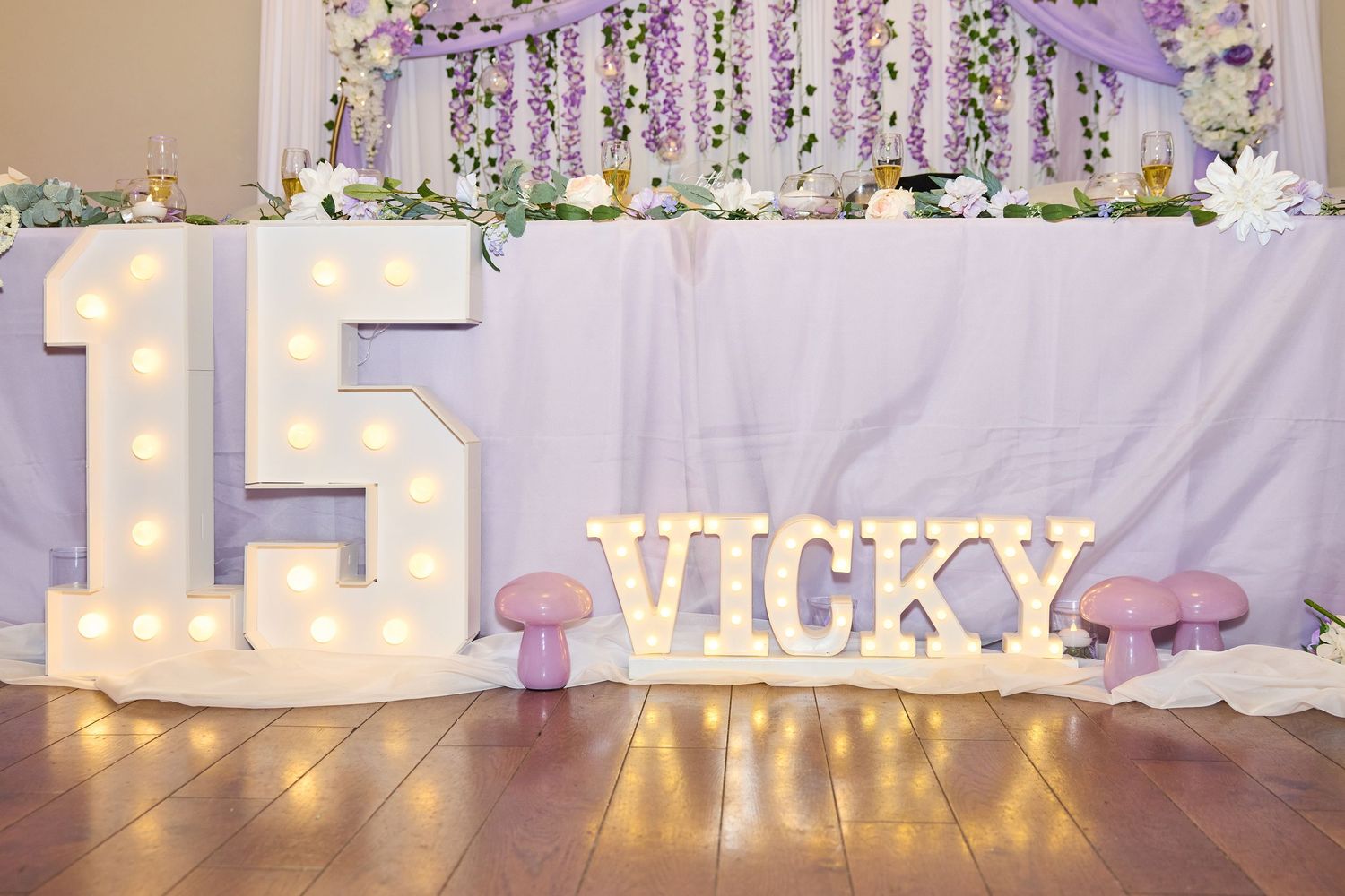 Elegant wedding head table setup with illuminated letters and purple floral decor against white backdrop.