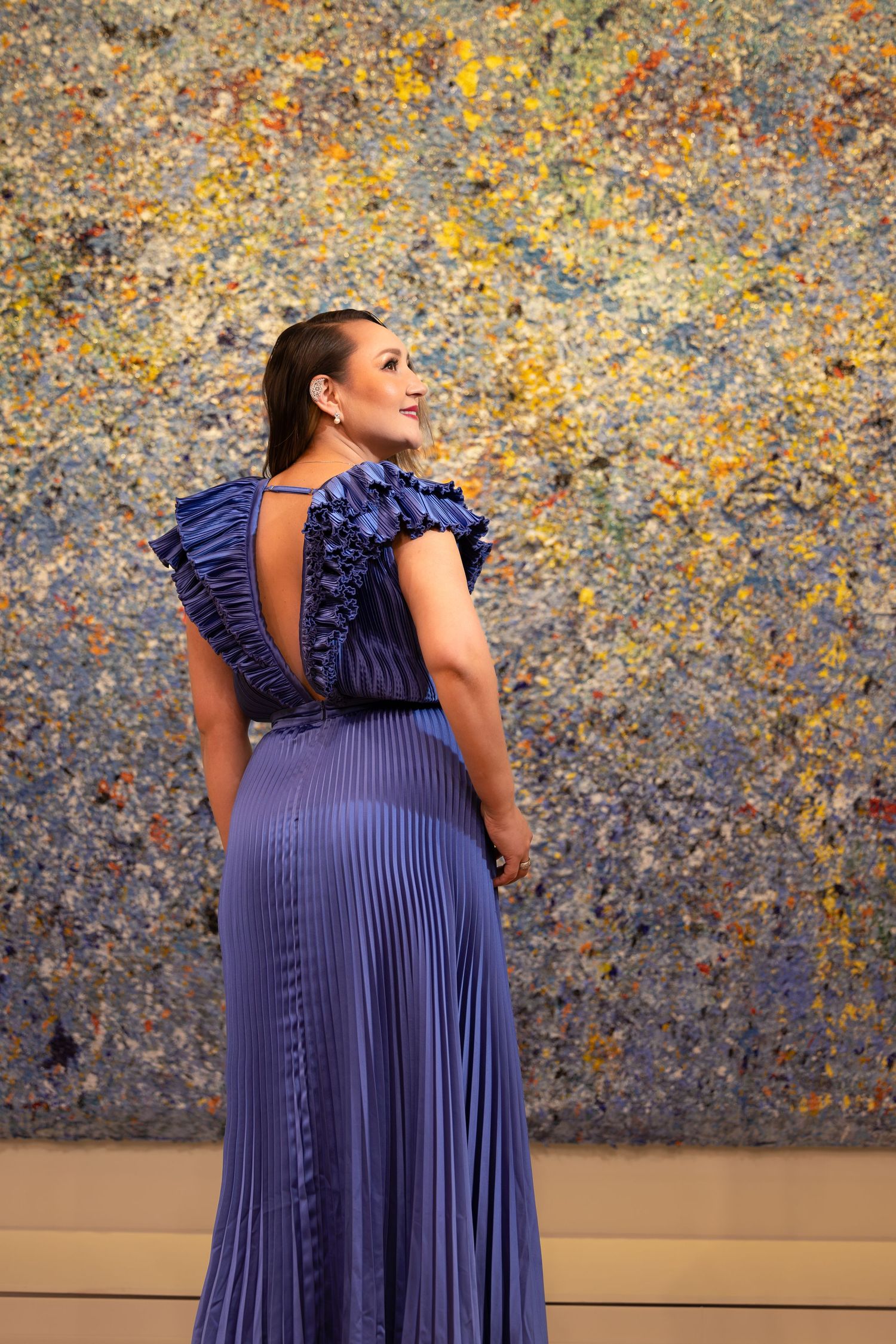 Back view of pleated navy gown with ruffled shoulders against vibrant abstract wall art.