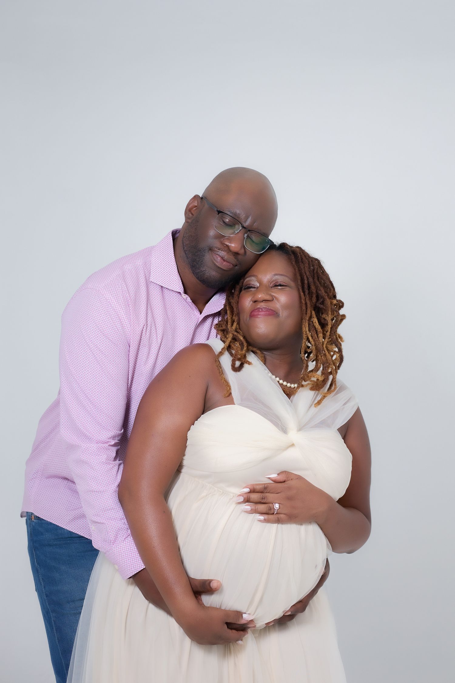A couple embraces tenderly during a maternity photo shoot, the woman in white dress cradling her baby bump.
