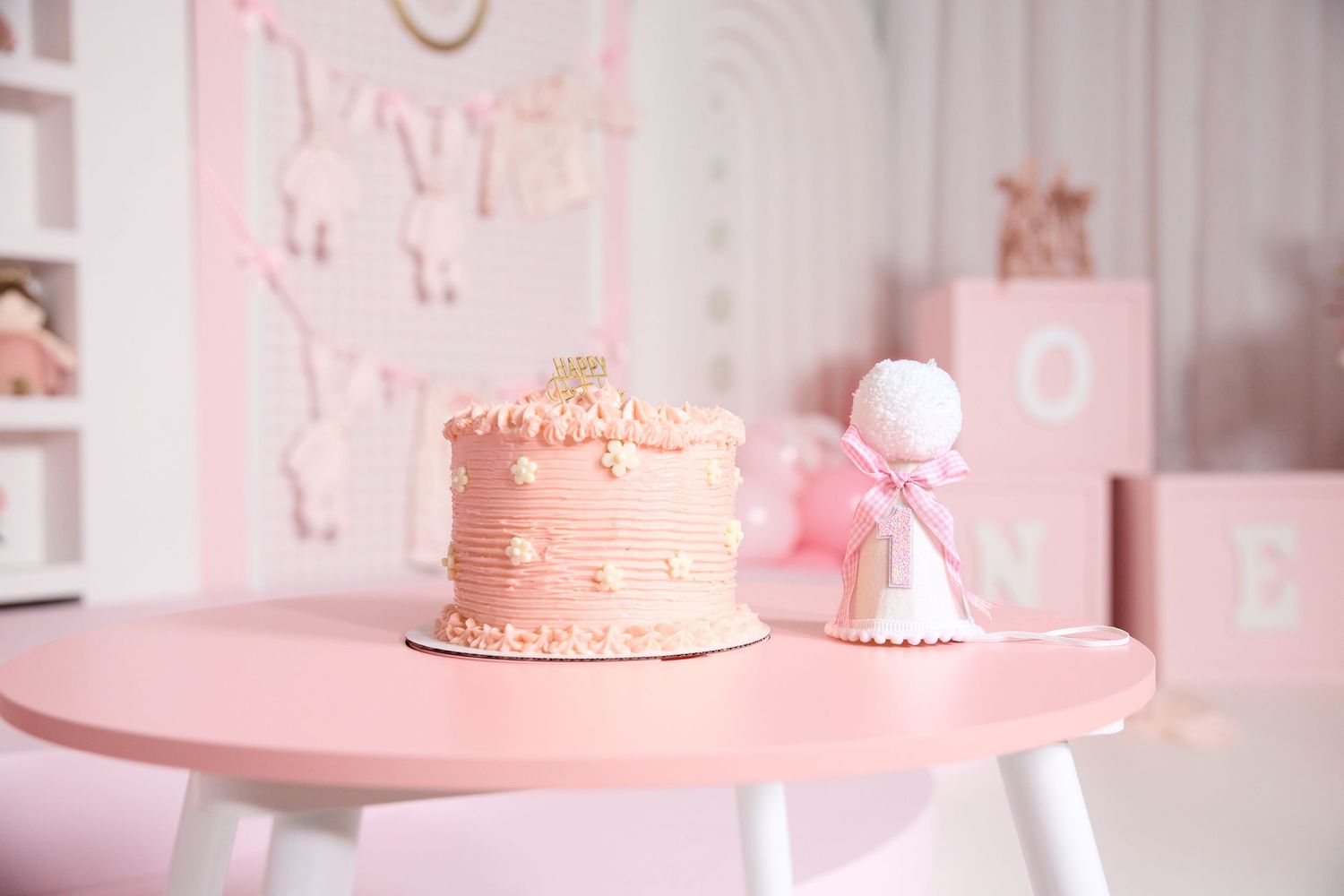 Pink first birthday cake with pearl decorations on a pink table with a small figurine and ONE block in a pastel decorated room.