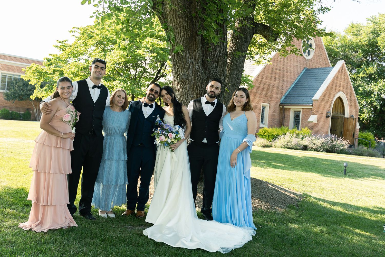 A wedding party in formal attire poses outdoors on a sunny day with a small church and green trees in the background.