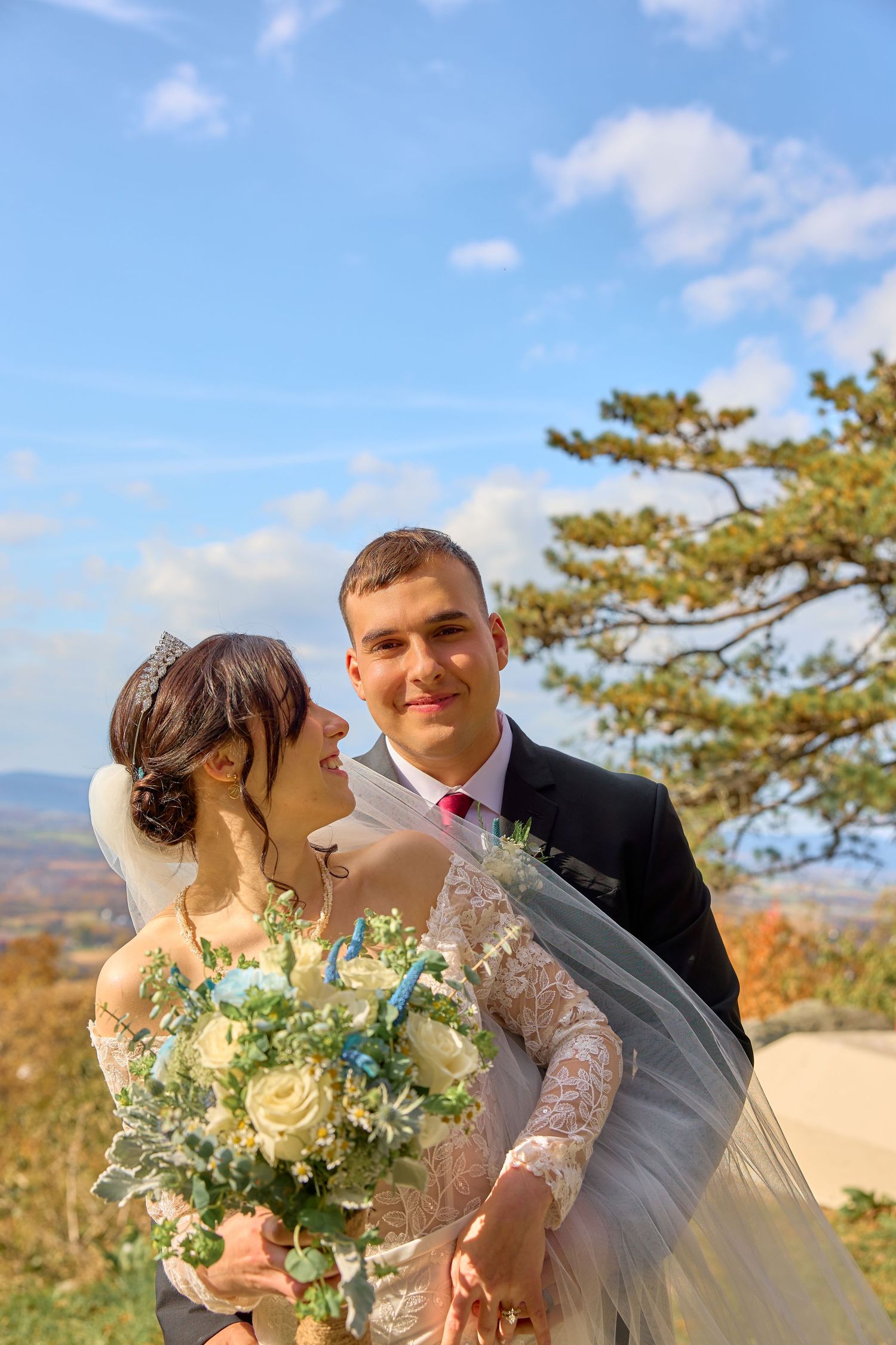 Newlyweds embracing outdoors with mountain scenery and pine trees in the background.