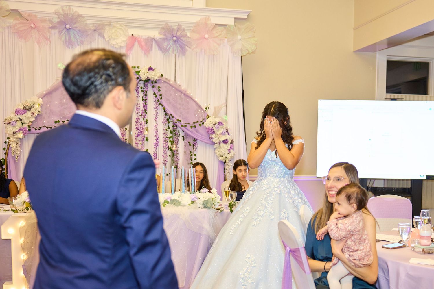 Guests dance and celebrate at an elegant wedding reception decorated with purple and white floral arrangements.