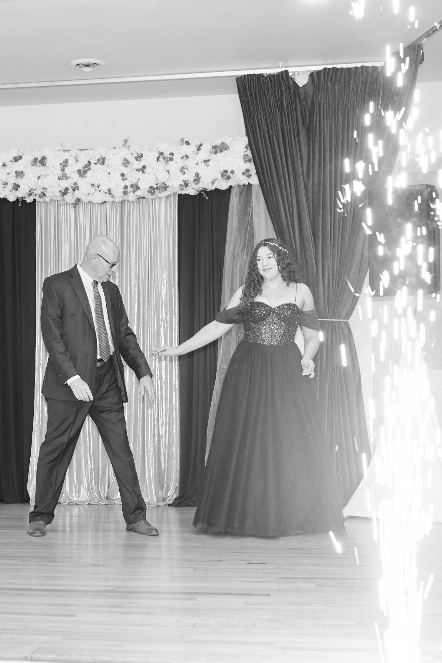 People dancing in formal attire at an event venue with sparkling lights and white curtains in the background.