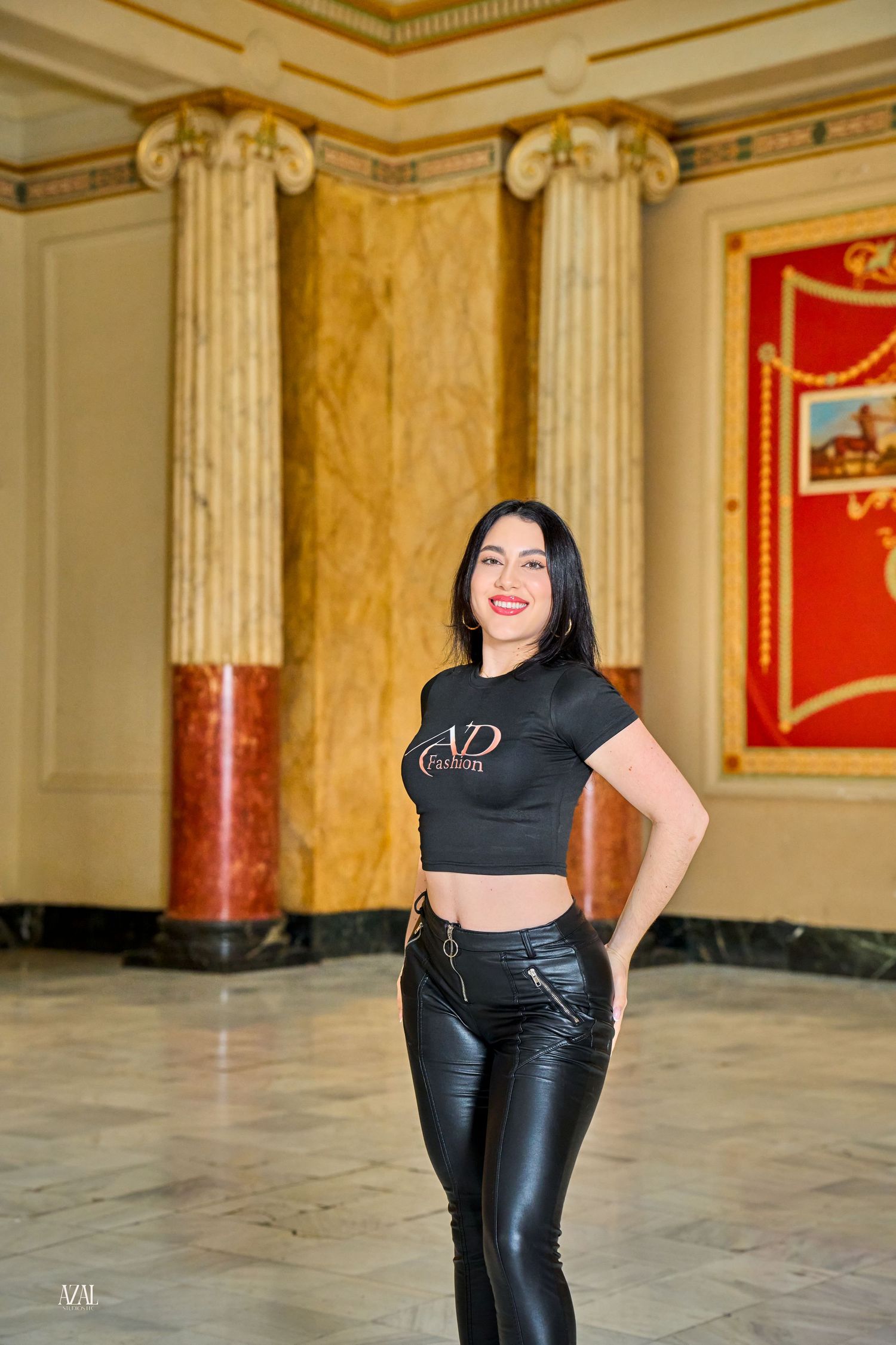 Woman in black VD Fashion crop top and leather pants smiles in grand hall with marble columns.