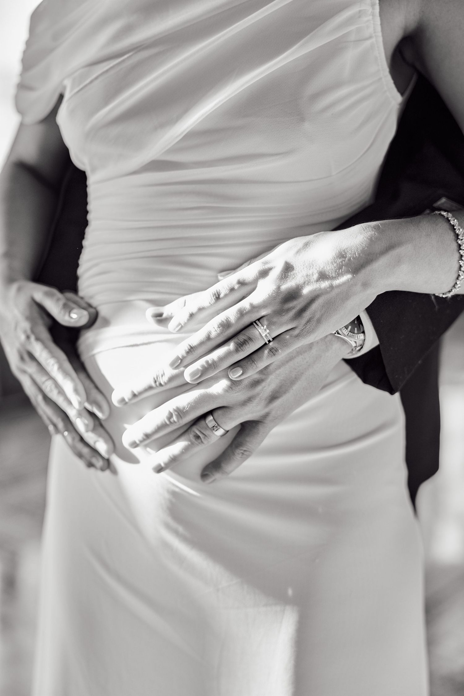 Black and white photo of a pregnant woman in elegant dress with partner's hands cradling her baby bump, wearing rings.