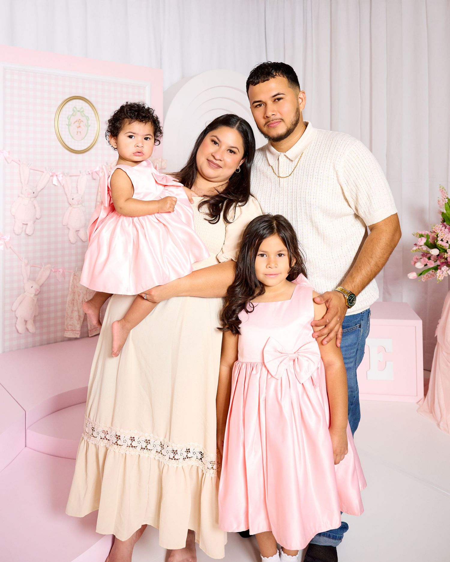 Family of four posing in pink studio, mother holding toddler in pink dress, father and older daughter smiling warmly.