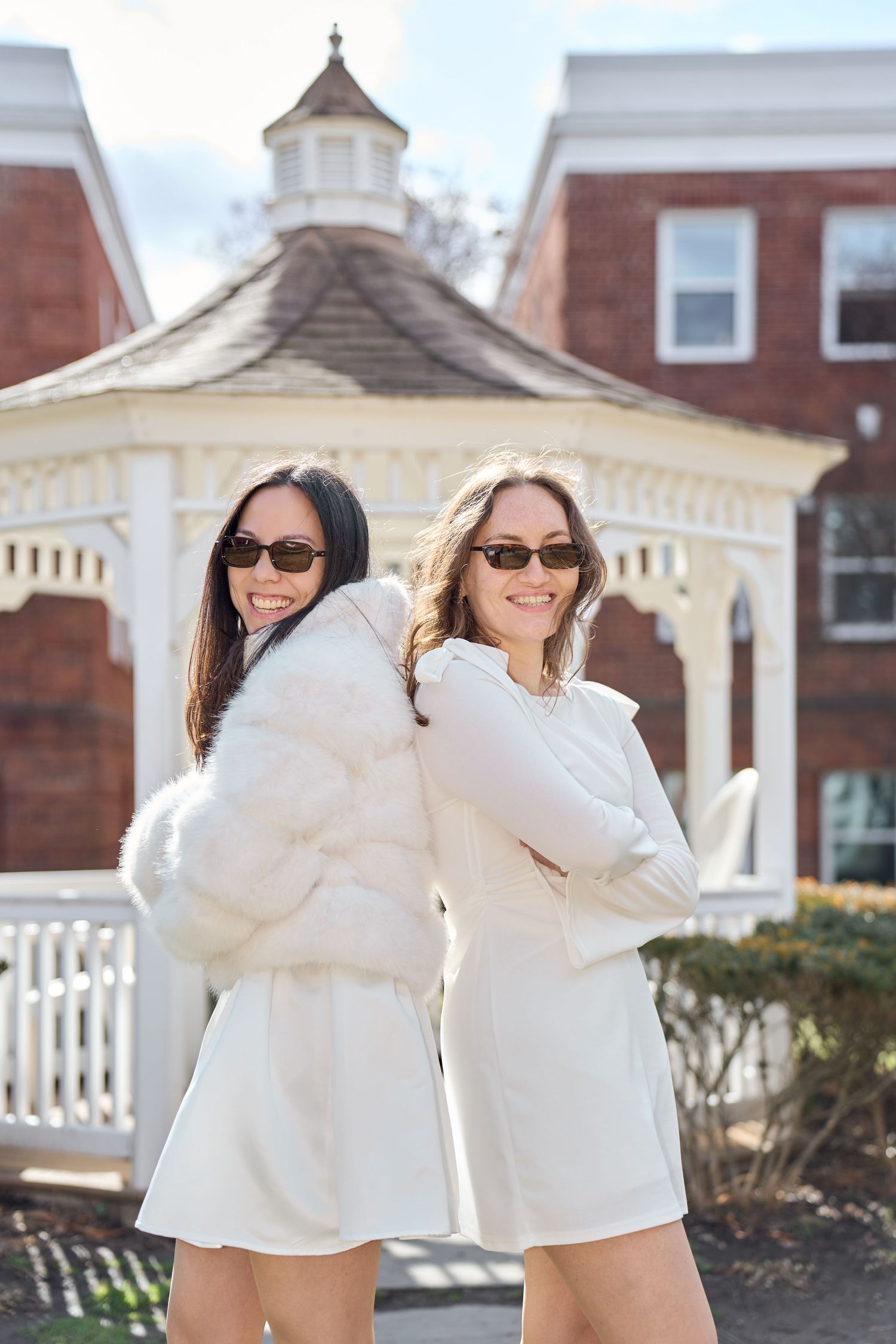 Two people in matching white outfits pose playfully outdoors with a classic brick building and gazebo in the background.