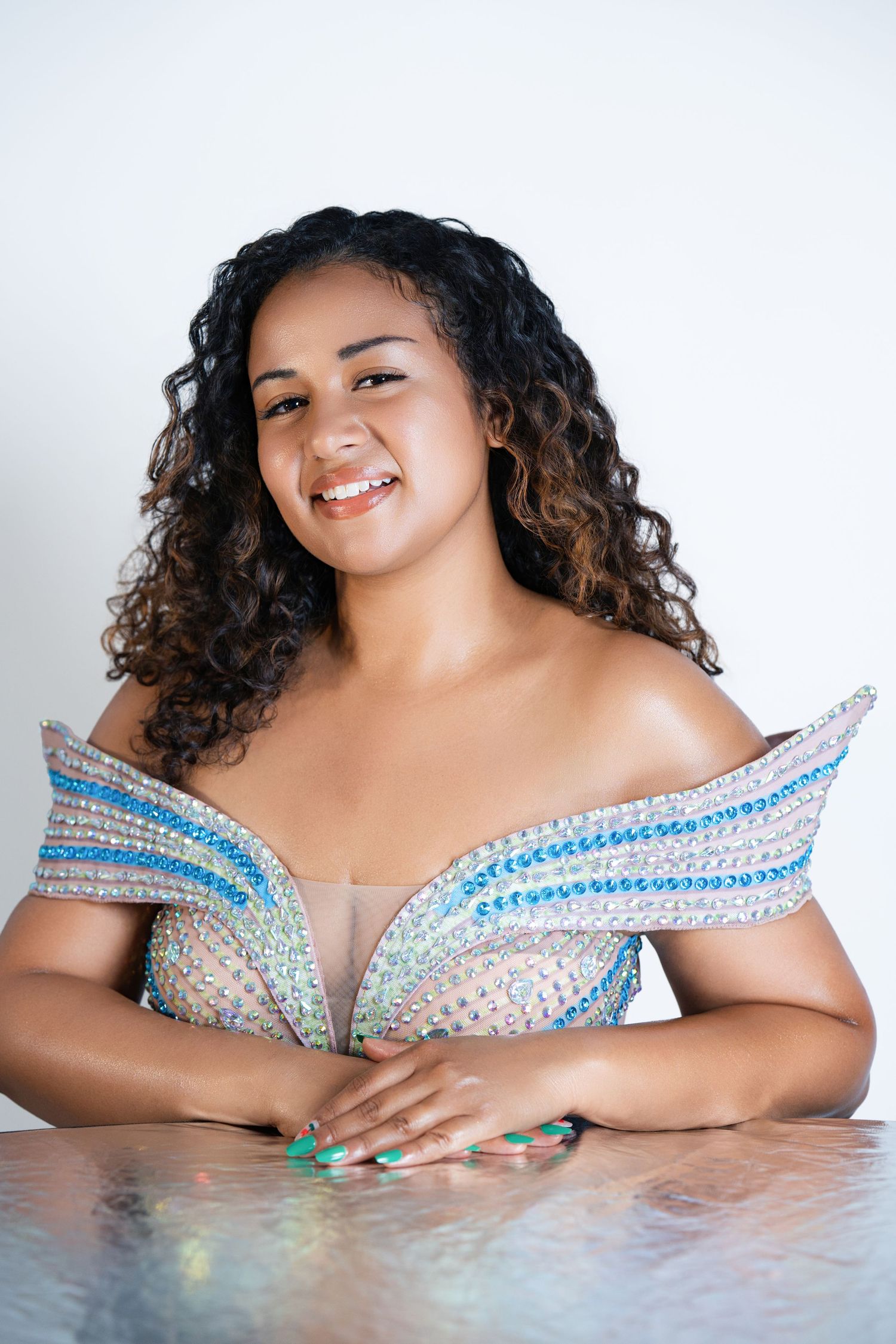 Smiling young woman with curly hair wearing a sparkly off-shoulder gown, posing at a reflective table.