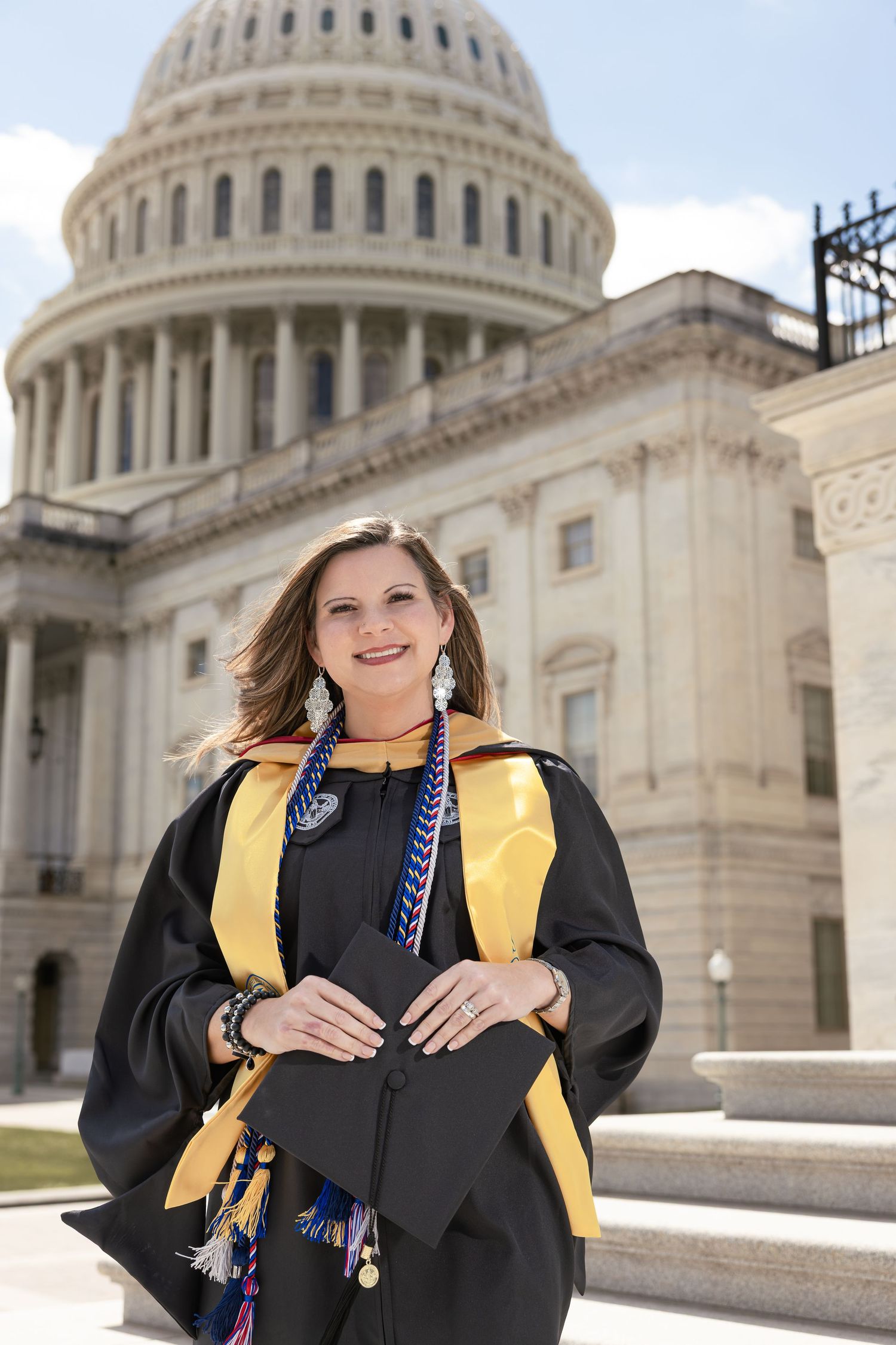 Graduate poses in cap and gown with gold stole in front of the United States Capitol building on a sunny day.