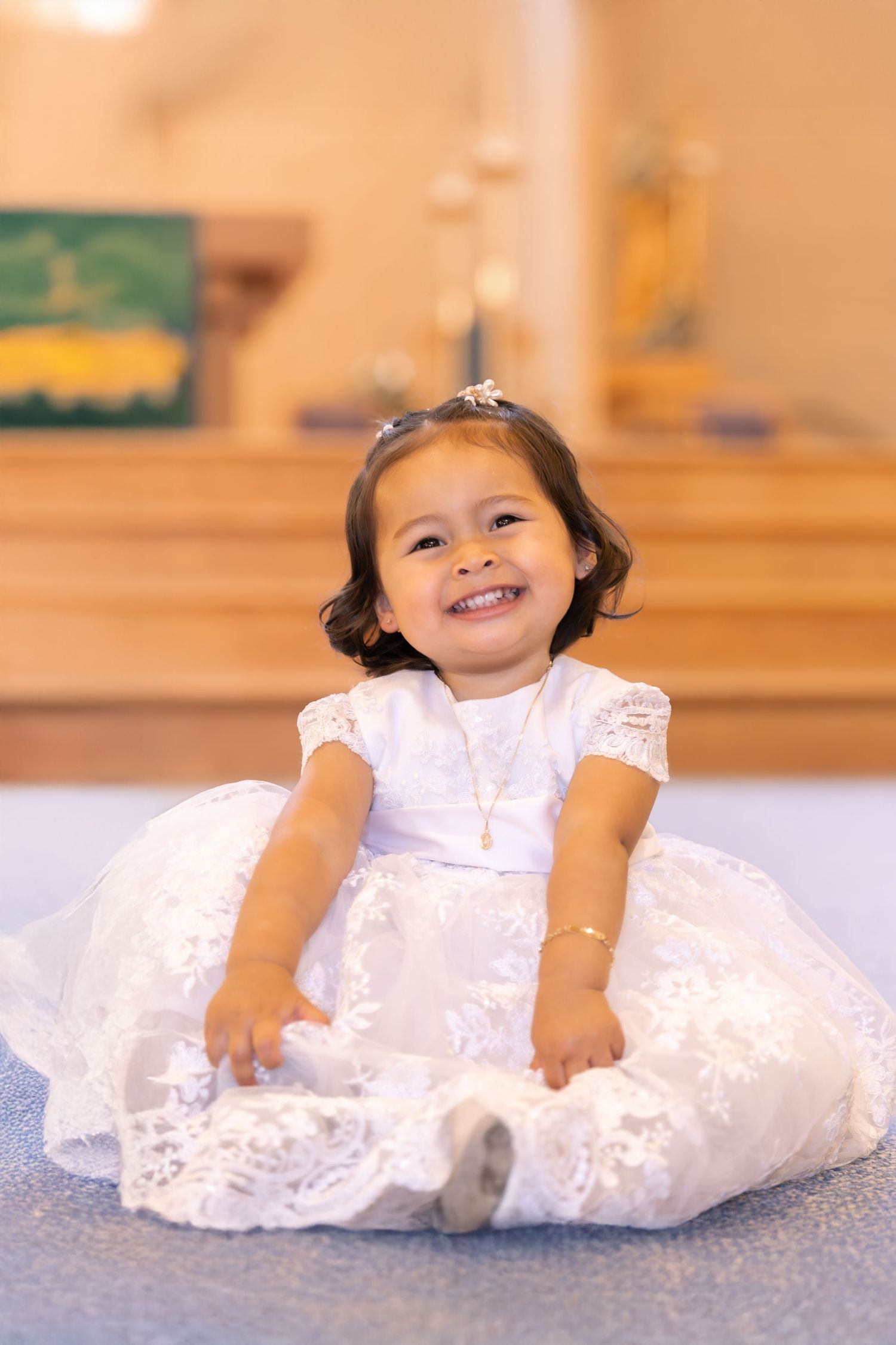 A toddler in a white lace dress sits on blue carpet while smiling brightly.