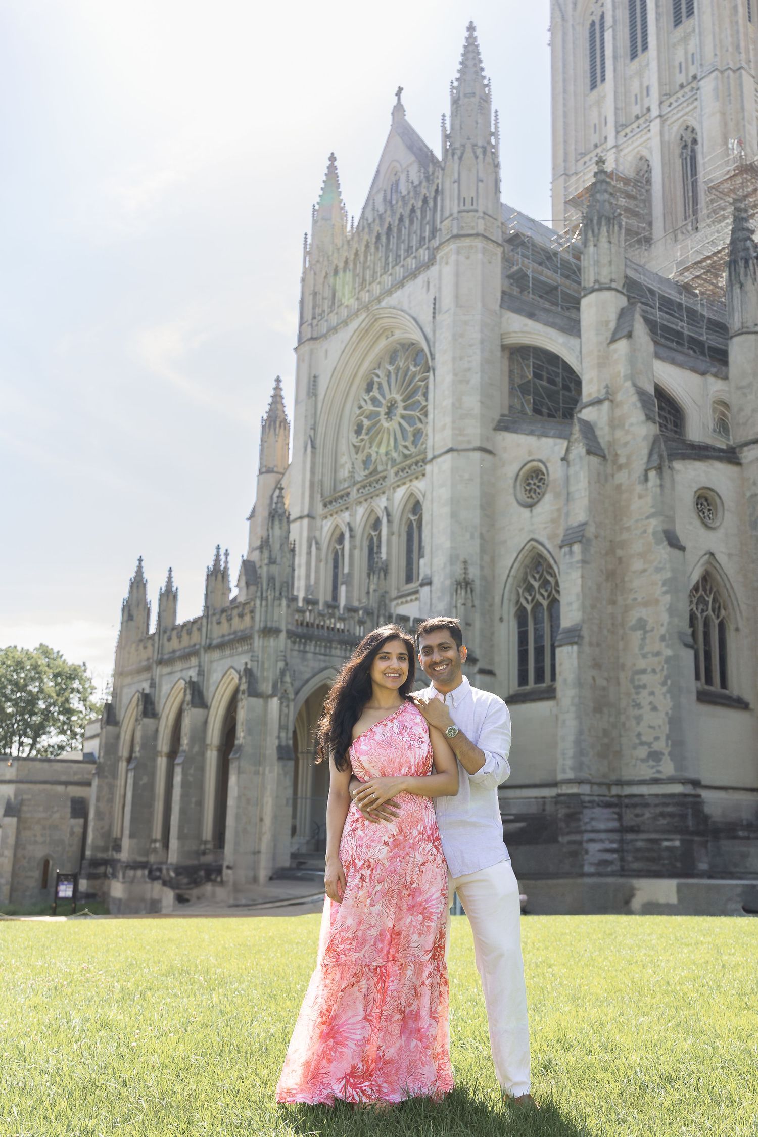 A couple poses together on a grassy lawn in front of a majestic Gothic cathedral with ornate stone architecture.