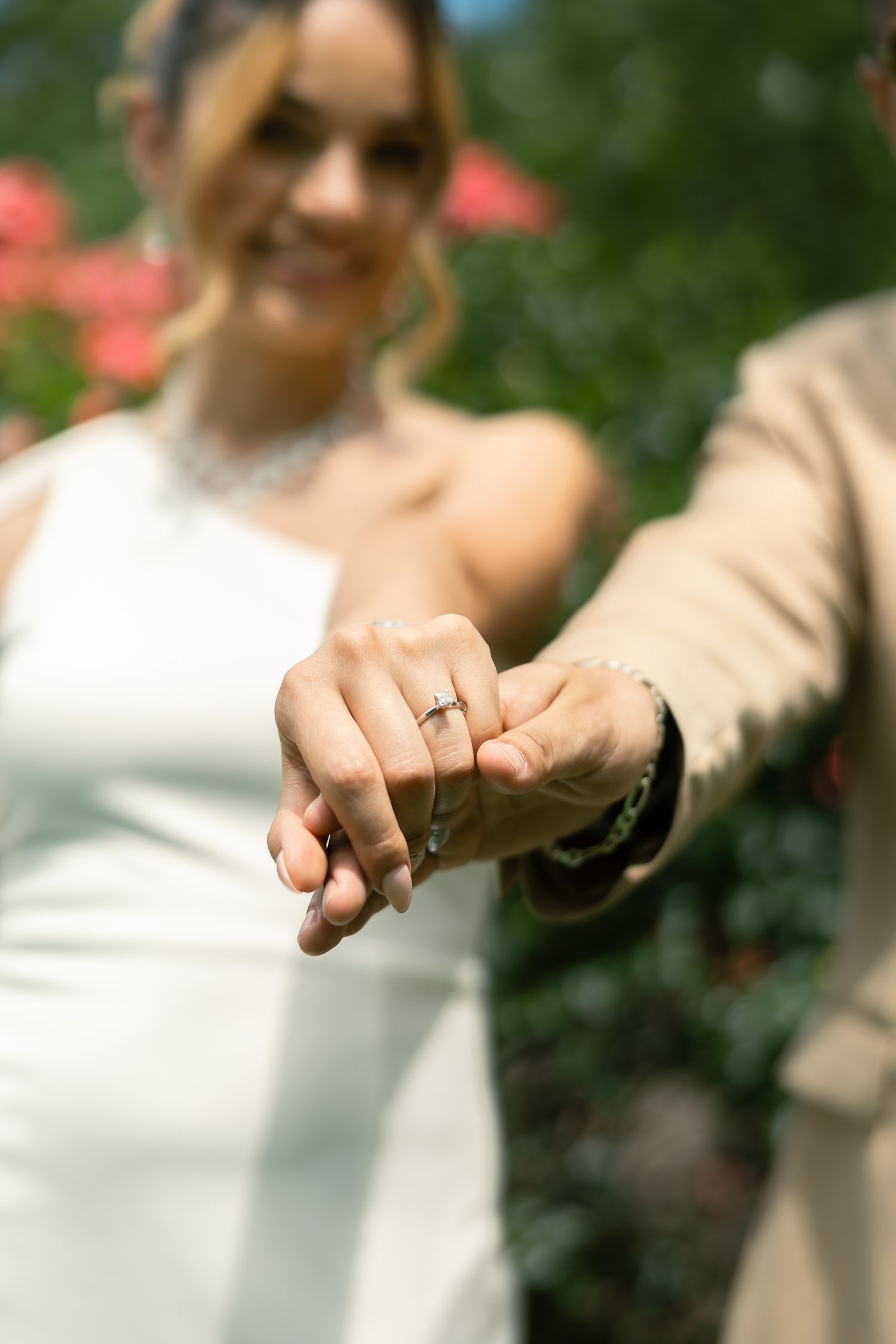 Close-up shot of intertwined hands showing off a wedding ring against a blurred floral background.