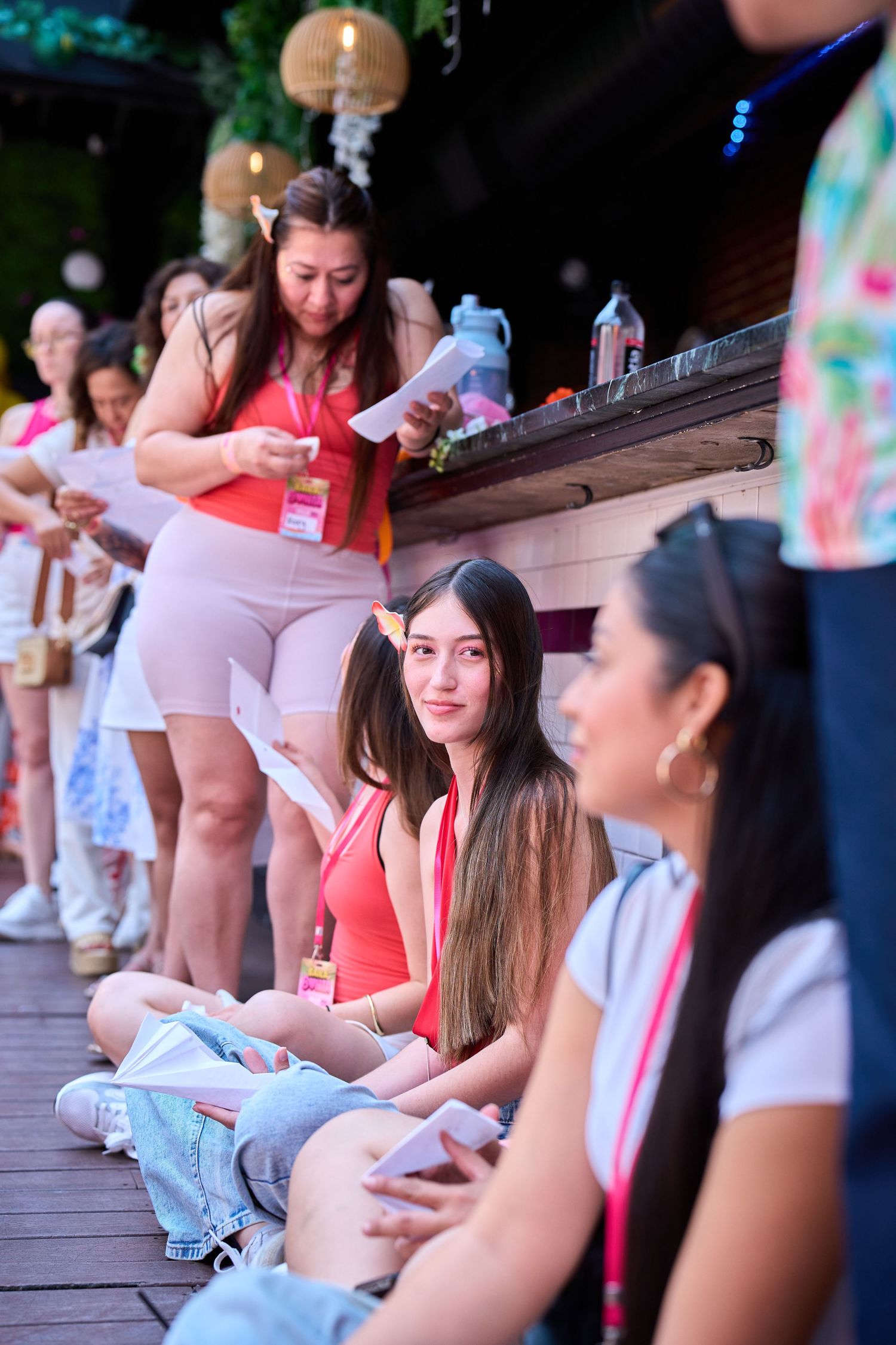 Young women sitting and standing on a wooden deck at an outdoor event, wearing colorful outfits and lanyards.