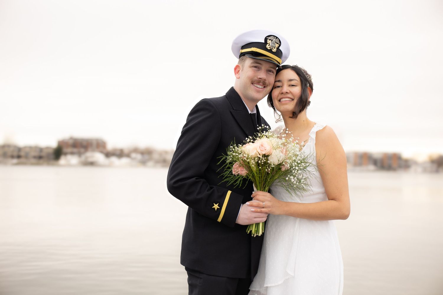 A joyful wedding couple poses by the water, with the groom in naval uniform.