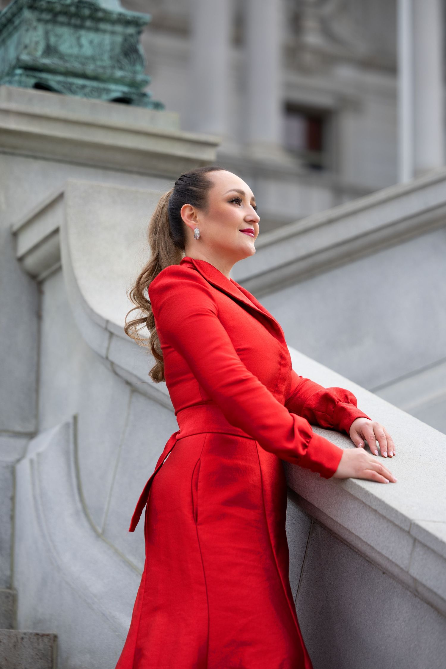 Woman in fitted red coat dress poses confidently against classical architecture.