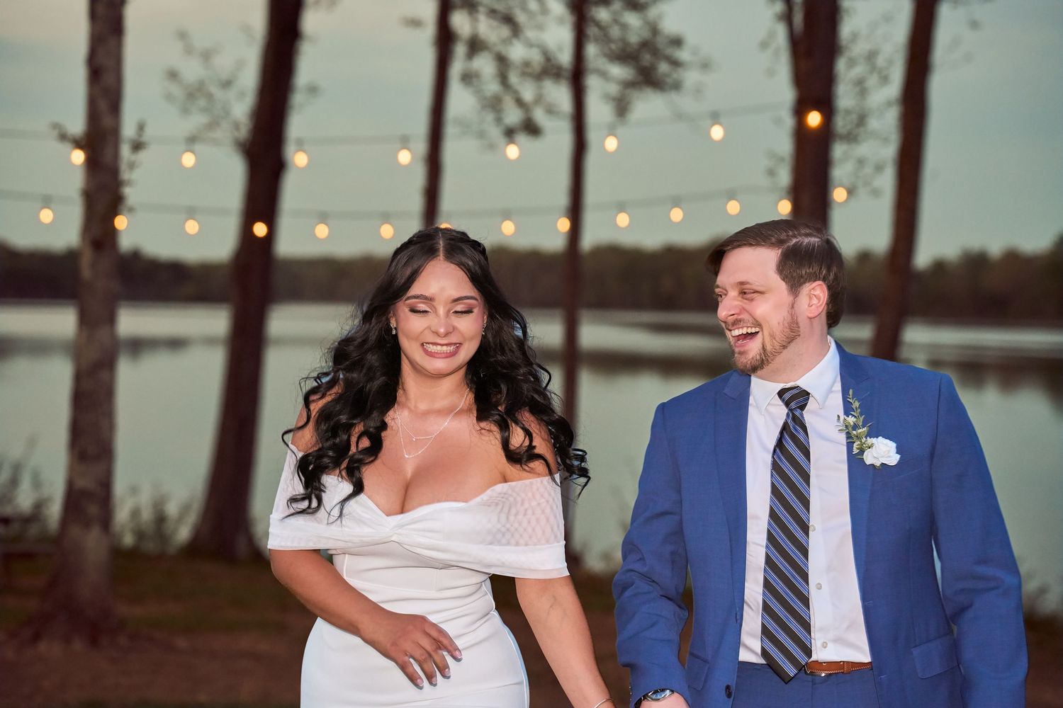 Bride in white off-shoulder gown and groom in blue suit hold hands outdoors near lake with string lights at dusk.