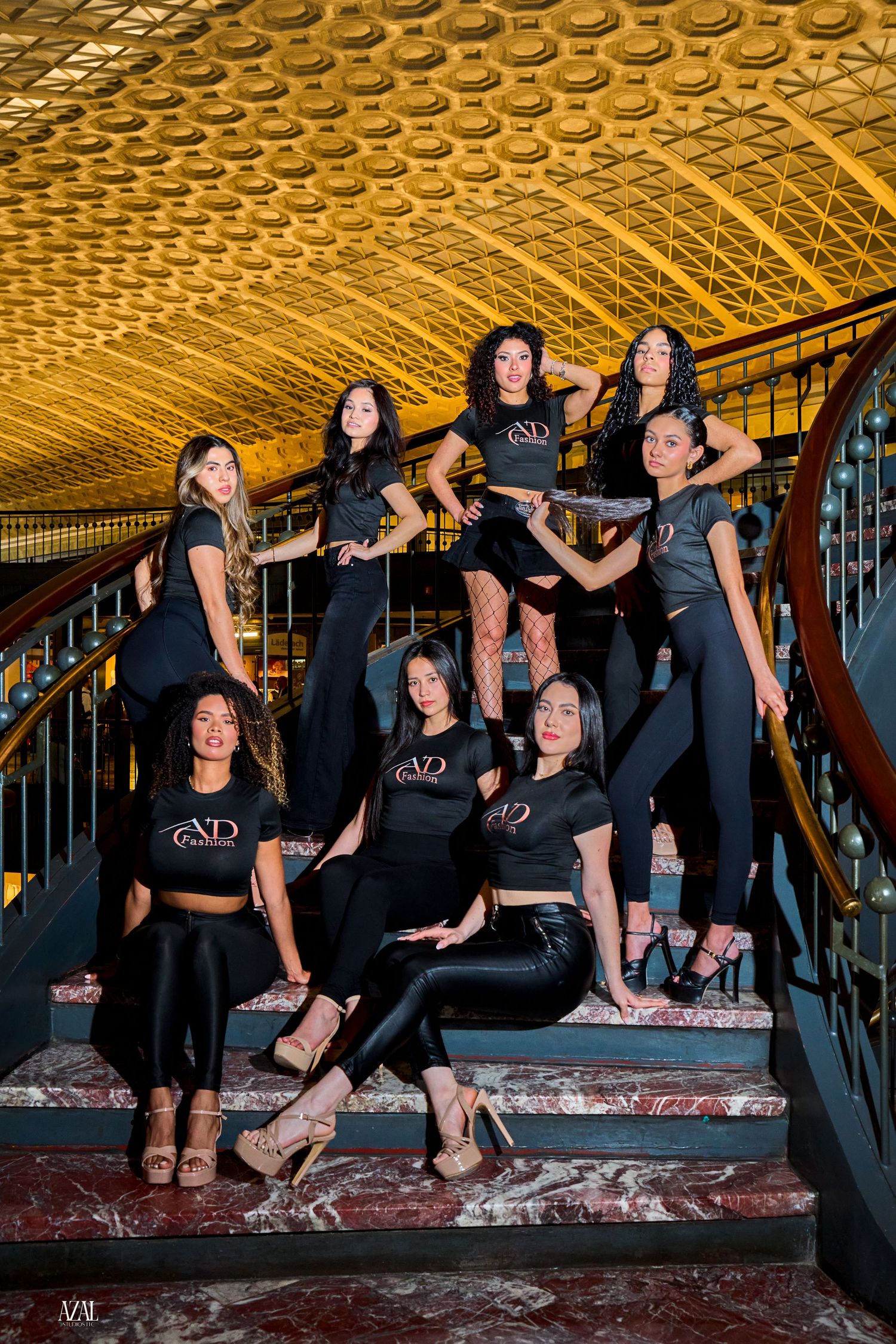 Group of women in matching black ND branded outfits posing on ornate marble stairs in elegant golden venue.