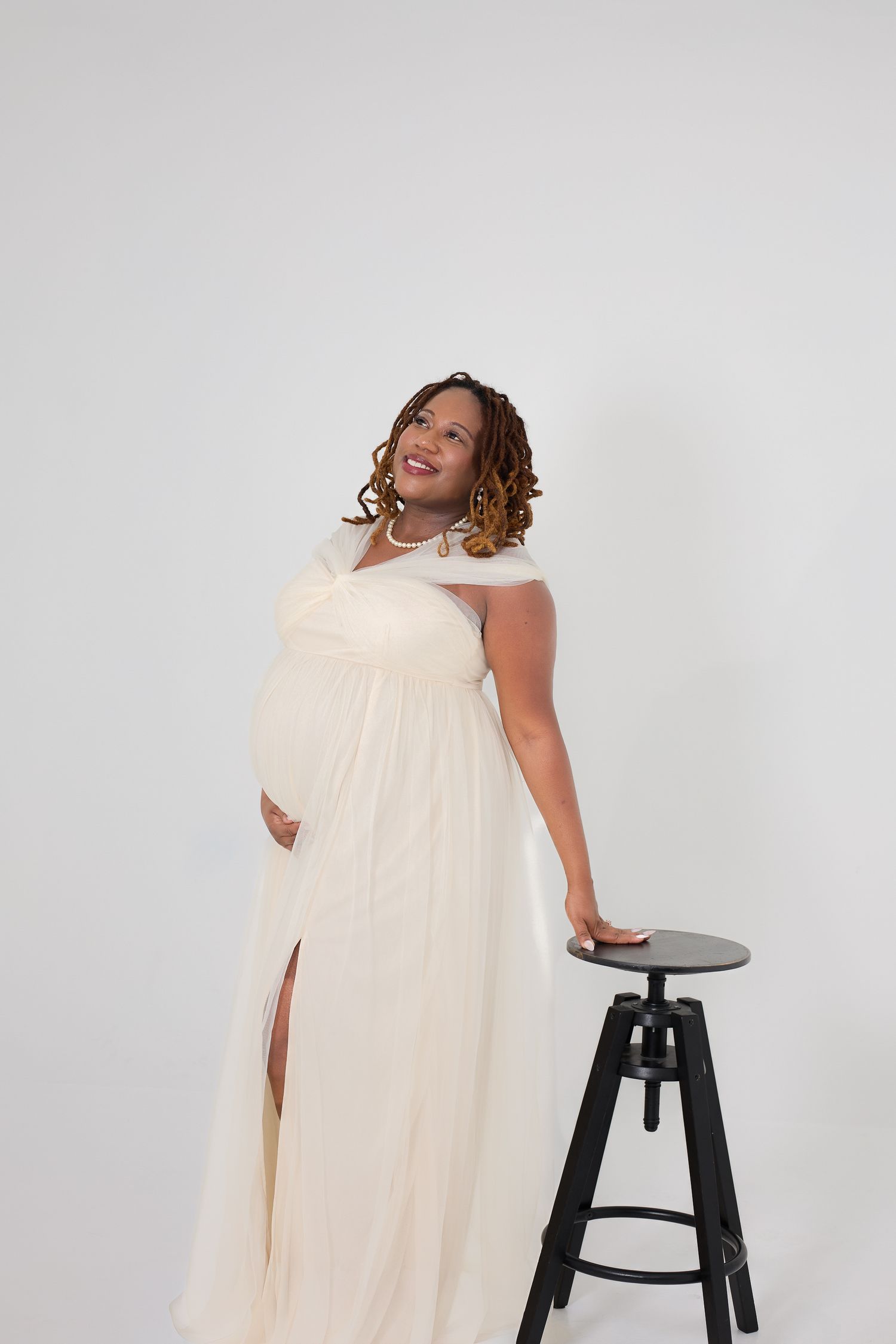 Pregnant woman in elegant cream maternity gown posing beside a black stool against white background.