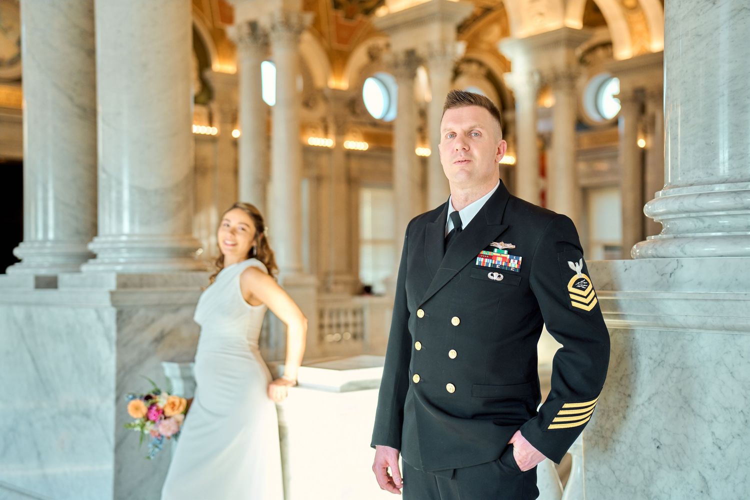 Navy officer in dress uniform stands with bride holding bouquet in elegant marble hall with columns.