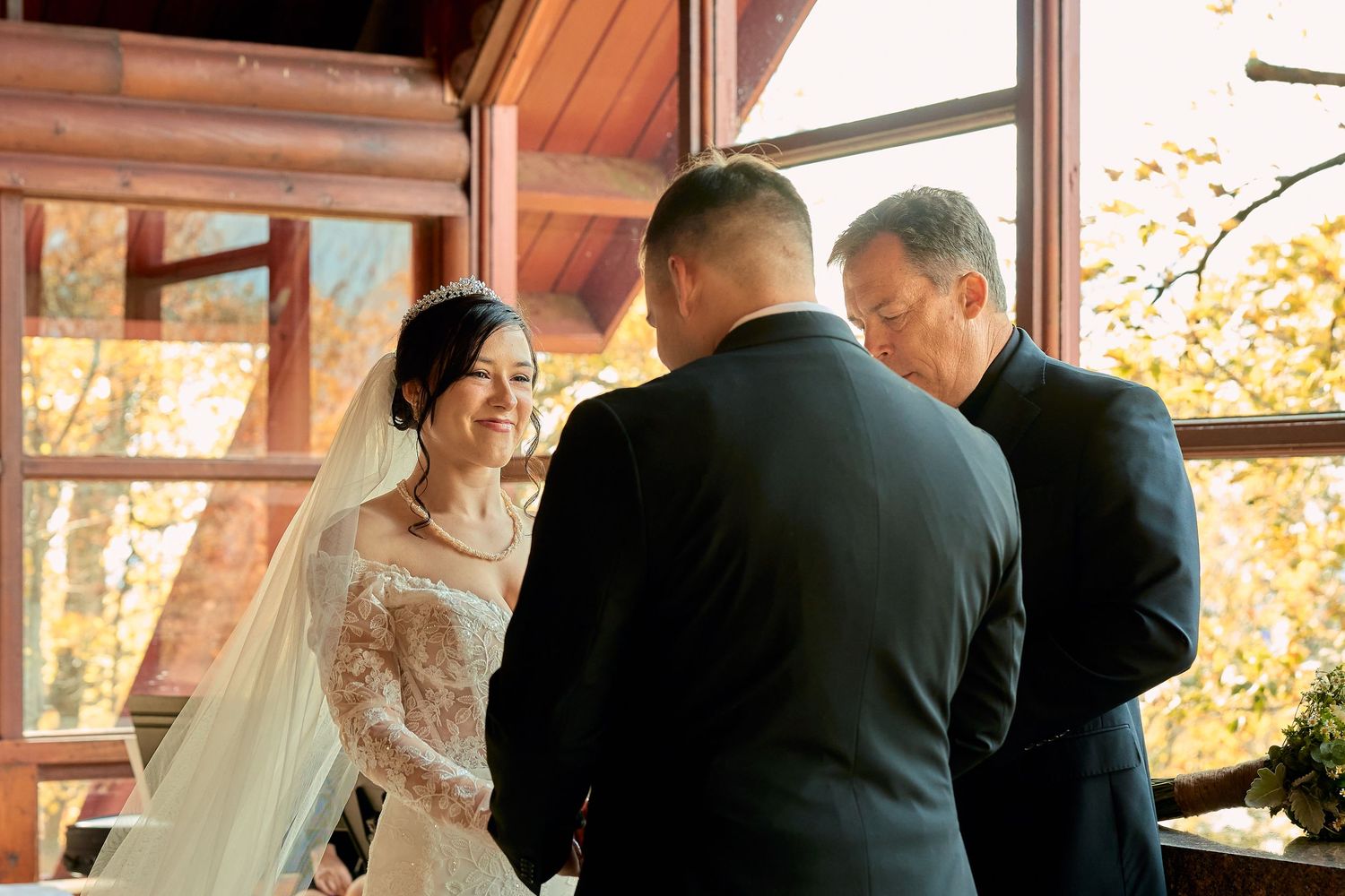 Wedding ceremony in a rustic venue with wooden beams and floor-to-ceiling windows showing fall foliage.