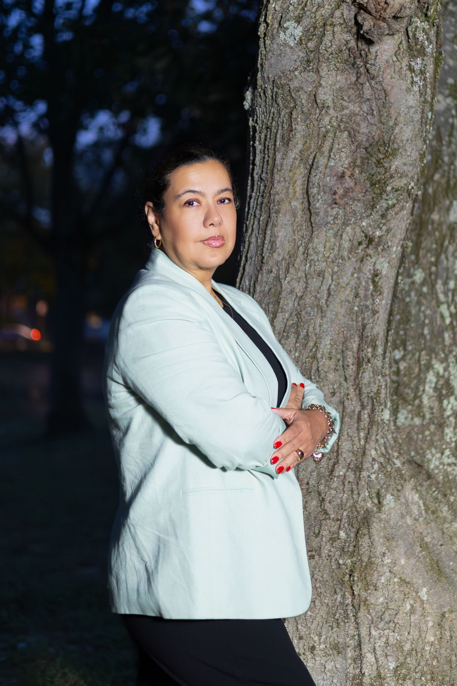 A person in a white winter coat poses against a tree trunk in an outdoor evening setting.