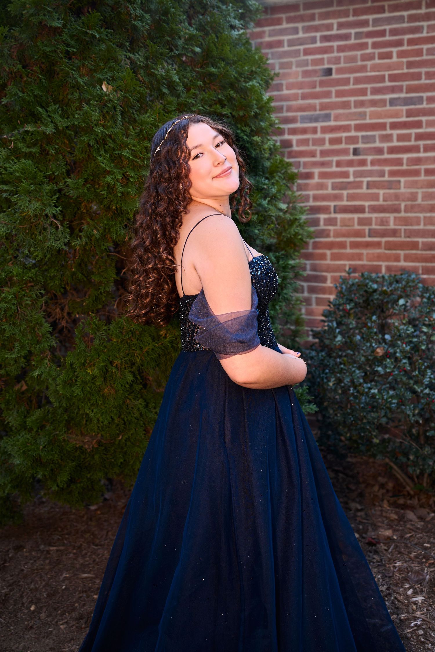 A person in a navy blue formal gown poses against a brick wall and greenery in elegant evening attire.