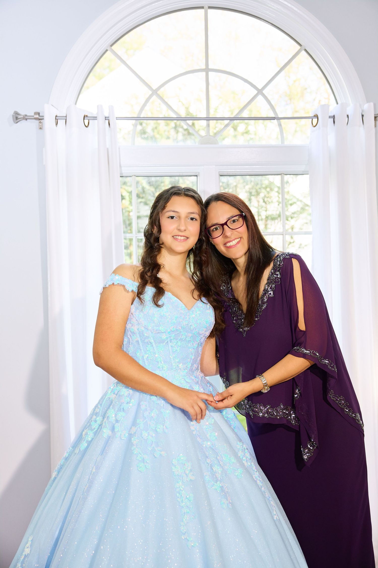 Two people pose together in formal attire, one wearing a light blue ballgown and one in a purple dress by an arched window.