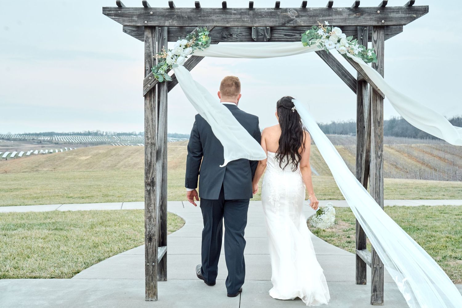 A couple walks along a white wooden pergola path with rolling hills and countryside in the background.