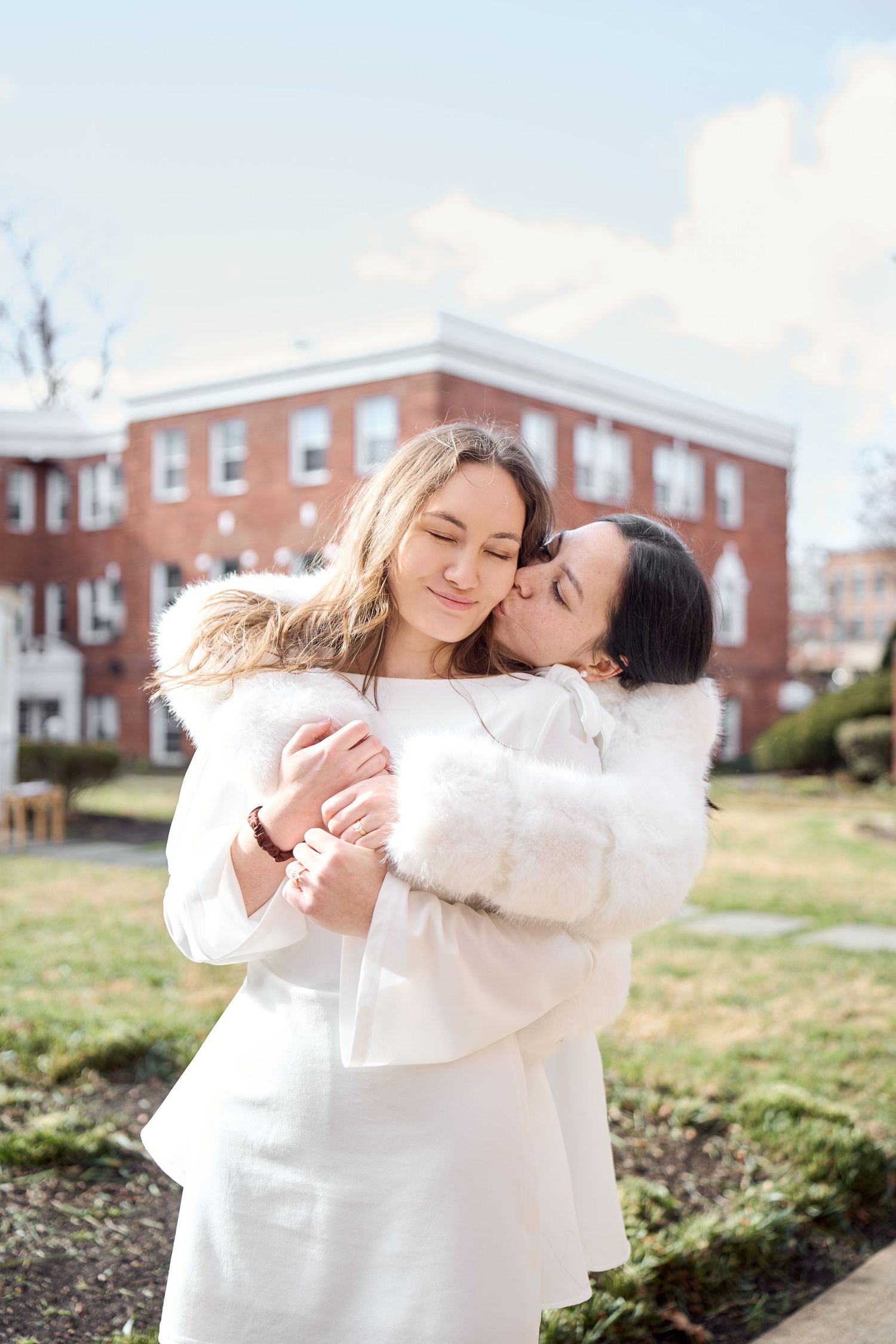 Two people in white outfits share a tender moment embracing outdoors with brick buildings in the background.