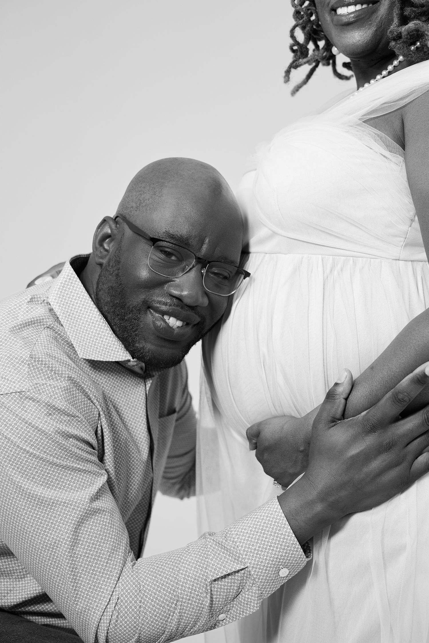 Black and white maternity photo of a smiling man with glasses leaning against pregnant womans belly.