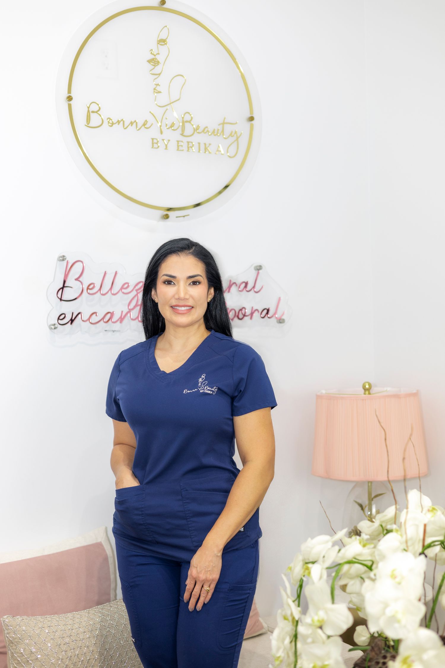 Healthcare worker in navy blue scrubs stands in front of decorative wall signs and floral arrangement in medical office setting.