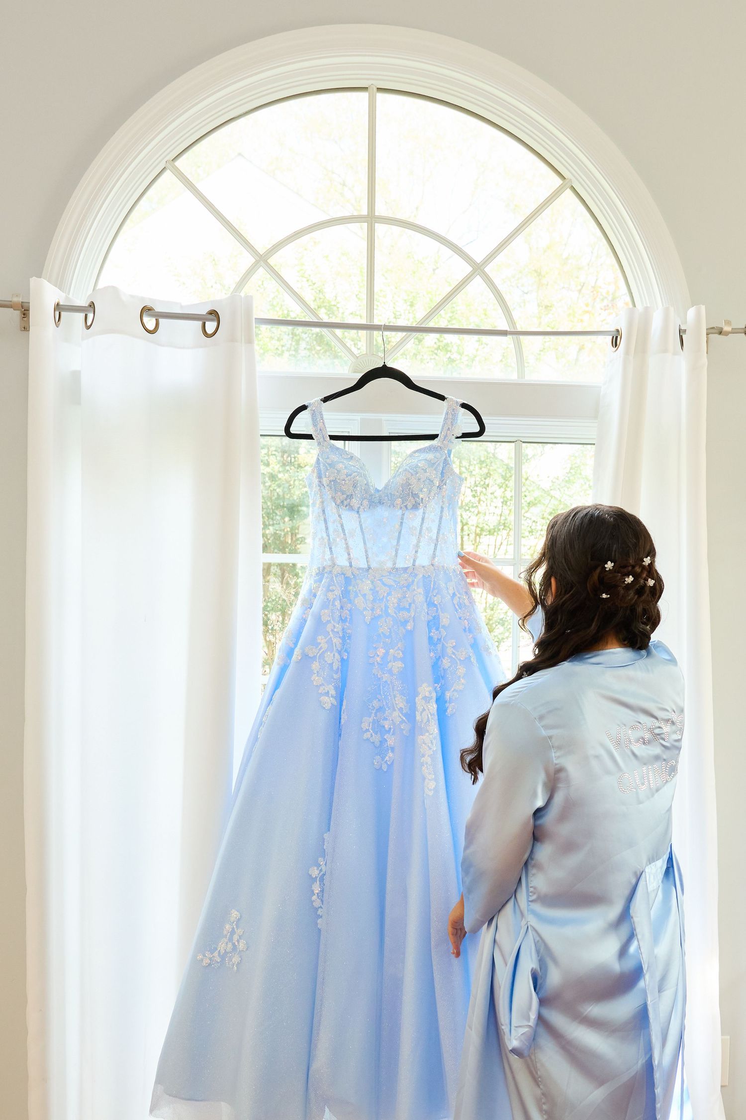 Light blue formal dresses with delicate embellishments hanging on display in a bright white room with arched windows.