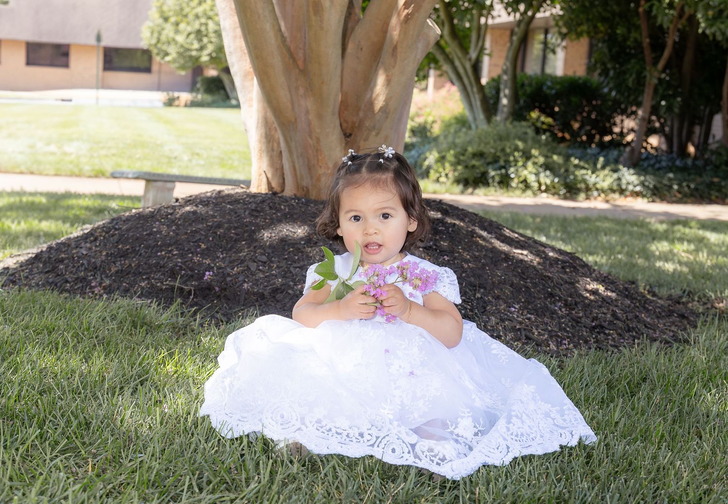 A small toddler in an elegant white lace dress sits on green grass in the sunshine.