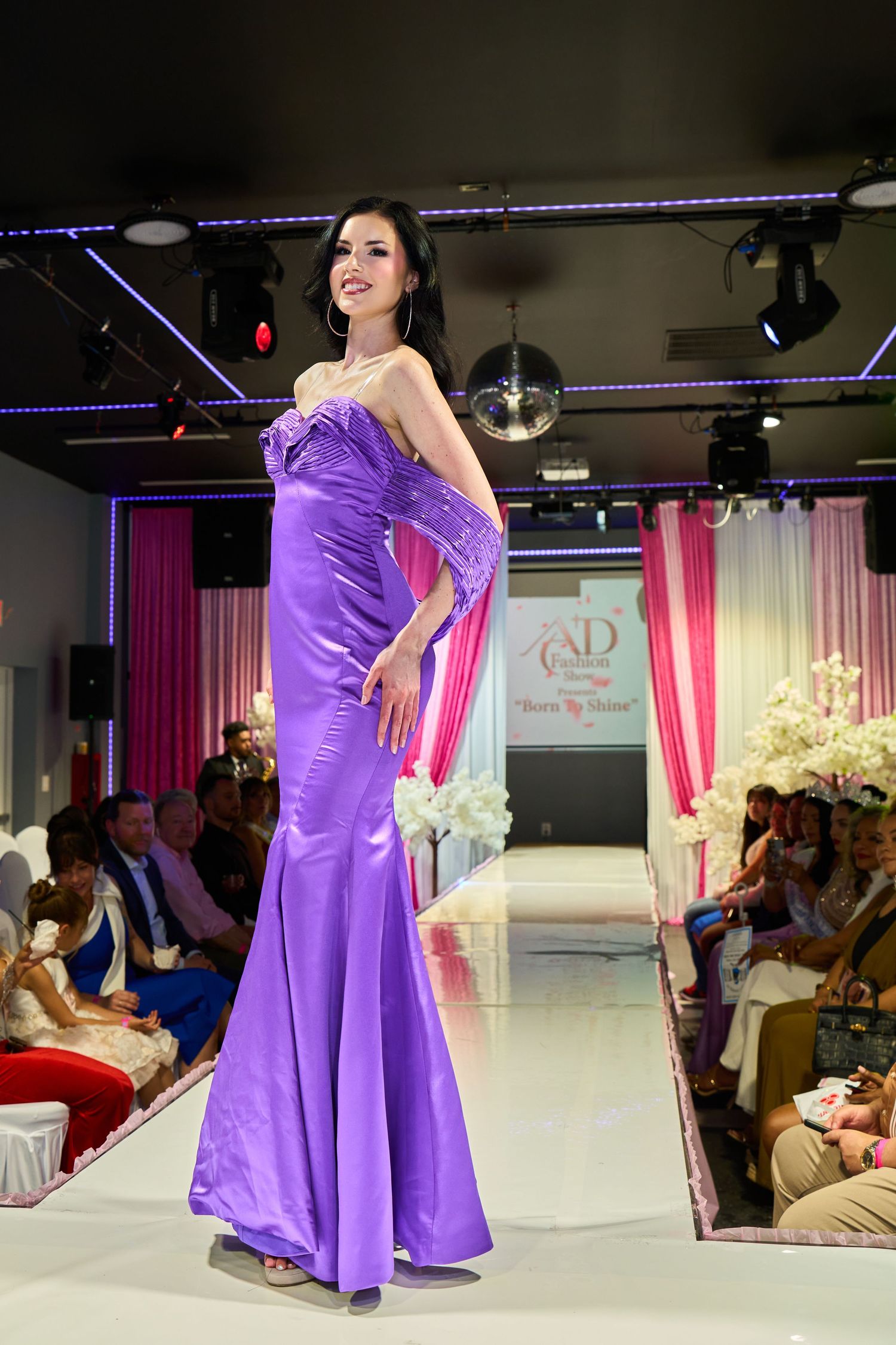 Model walks runway in elegant purple satin mermaid gown at indoor fashion show.