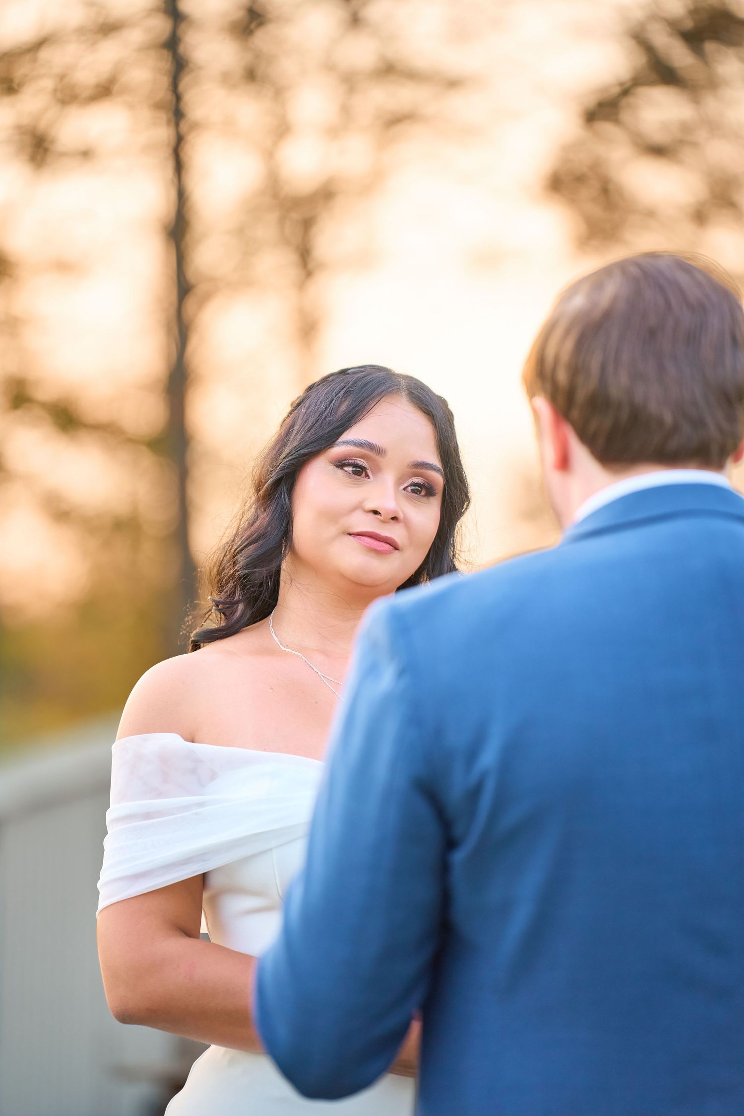 Bride in off-shoulder white gown gazes at groom in blue suit during golden hour outdoor wedding ceremony.